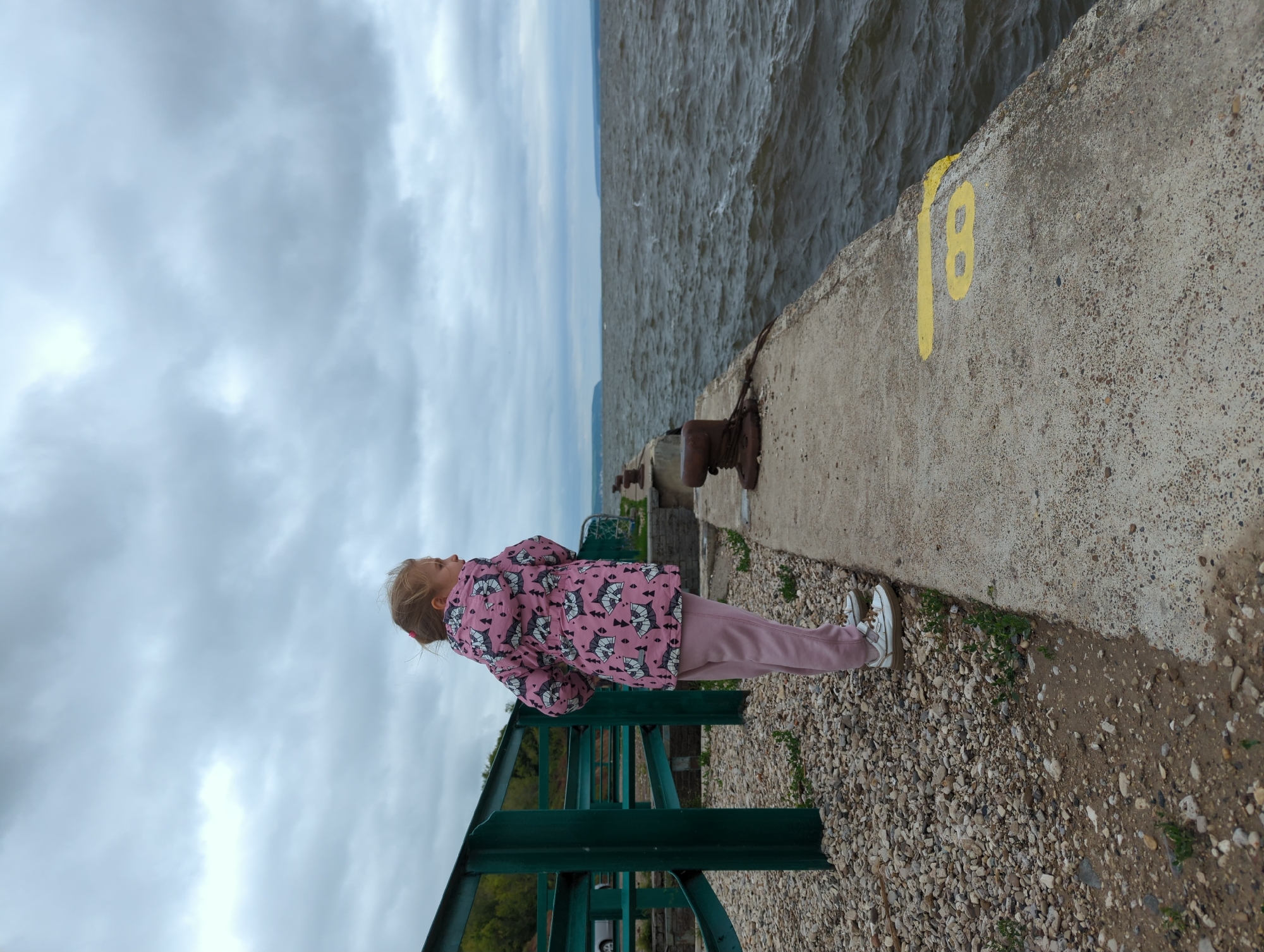 A young girl stands on a concrete pier, looking out at a choppy body of water. The girl is wearing a pink jacket with a patterned print and pink sweatpants, and white sneakers. She has blonde hair pulled back. The pier is made of concrete and is lined with dark metal bollards. To the left of the pier, there is a gravel area with some green metal structures. The sky is overcast with grey clouds. The location is Kamskoye Ustye, Russia, at the confluence of the Volga and Kama rivers.