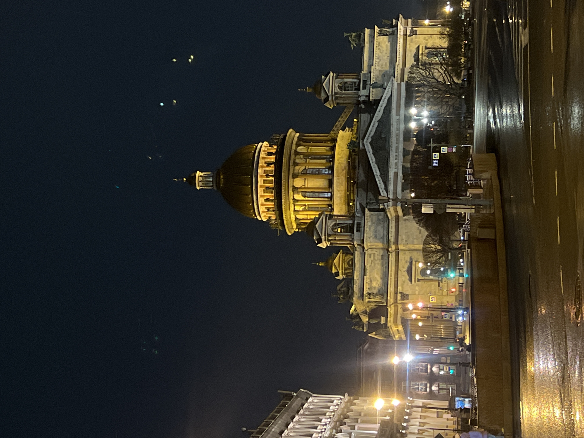 The image shows St. Isaac's Cathedral in Saint Petersburg, Russia, at night. The cathedral is illuminated, with its dome and facade visible. A wet road runs alongside the building. City lights are seen in the distance.