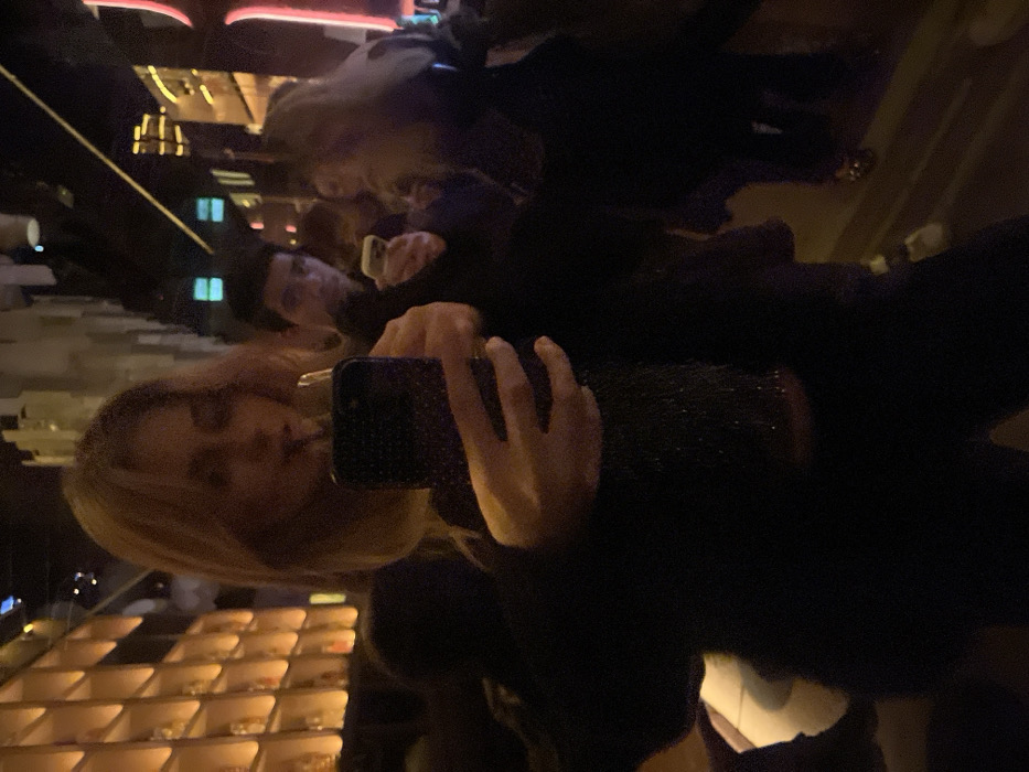 The image shows a woman with blonde hair holding a smartphone, seemingly taking a photo in a mirrored environment. Her reflection is visible multiple times, and another person is partially visible in the background. The setting appears to be a dimly lit interior, possibly a restaurant or bar, with decorative lighting and geometric patterns visible in the reflections. The overall image has a dark and distorted appearance.