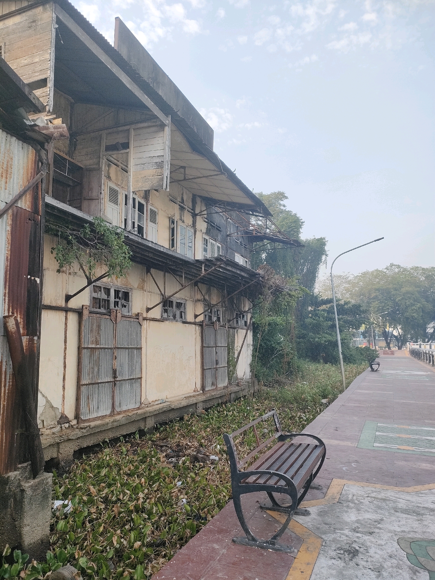 The image depicts a weathered, derelict building situated along a waterway in Banjarmasin, Indonesia. The building, likely an old warehouse or residential structure, shows significant decay with peeling paint, rusting corrugated metal siding, and broken windows. Overgrown vegetation spills from the building's lower levels and into the murky waterway, which is choked with water hyacinths and debris.
Adjacent to the building is a paved walkway, suggesting a public area or promenade. Several empty park benches are visible along the walkway, one in the foreground and another further in the distance. A lamppost stands near the walkway, its light fixture pointed upwards. In the background, lush trees and a hazy sky contribute to a slightly overcast atmosphere. The scene appears to be devoid of human activity at the moment the photograph was taken. The overall impression is one of neglect and the passage of time, set against the backdrop of a potentially bustling city now showing signs of urban decay.