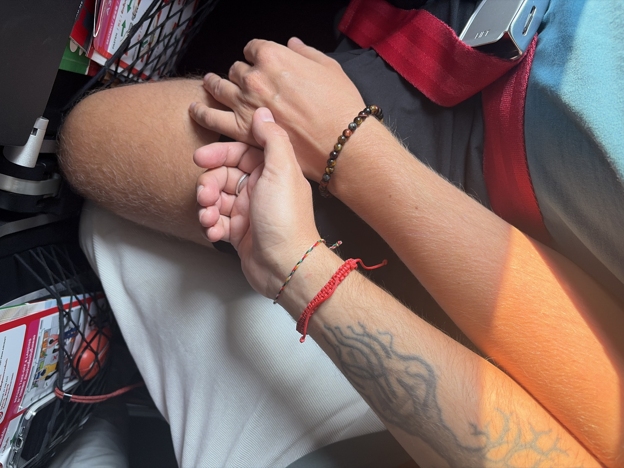 The image shows a close-up of two people seated next to each other, likely on an airplane. One person has a light-colored arm resting on the other person's leg which is covered in a white fabric. The person's hand has a silver ring on their ring finger. The other person has a darker complexion and a bracelet made of brown stones on their wrist, as well as a red woven bracelet on the other wrist, which also has a grey tattoo. A red seatbelt strap is visible in the background, as well as the airplane seat. The seat's back pocket contains reading materials and some small objects. Based on the text visible in the seat pocket, the flight is likely a Thai airline, the location is Chiang Mai City Municipality, Thailand.