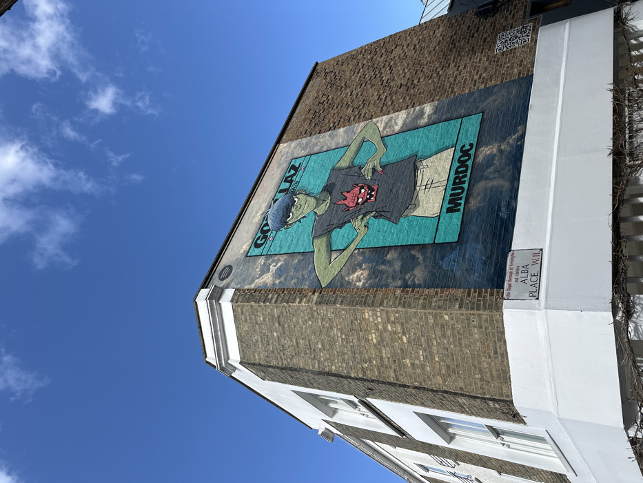 The image depicts the corner of a multi-story building located at Alba Place, W.11, within The Royal Borough of Kensington and Chelsea, London, United Kingdom. This location is identified by a white street sign affixed to the white-painted ground-floor base of the corner. The building's upper sections feature a brown brick facade with multiple white-framed sash windows visible on the left elevation and white trim along the corner edge.
A prominent mural covers a significant portion of the right-facing brick wall. This mural features Murdoc Niccals, a stylized character from the band Gorillaz. The character has green skin, spiky black hair, and wears a dark grey sleeveless t-shirt with a red horned demon design. He is depicted with hands on his hips, against a teal and cloudy sky background. The word "GORILLAZ" is lettered in white above the character, and "MURDOC" is lettered below. Above the mural, on the brickwork, a circular Spotify logo is visible. To the right of the mural, a black QR code is affixed to the brick wall at a lower height. The sky above the building is clear blue with scattered white cumulus clouds. No individuals are present in the visible scene.