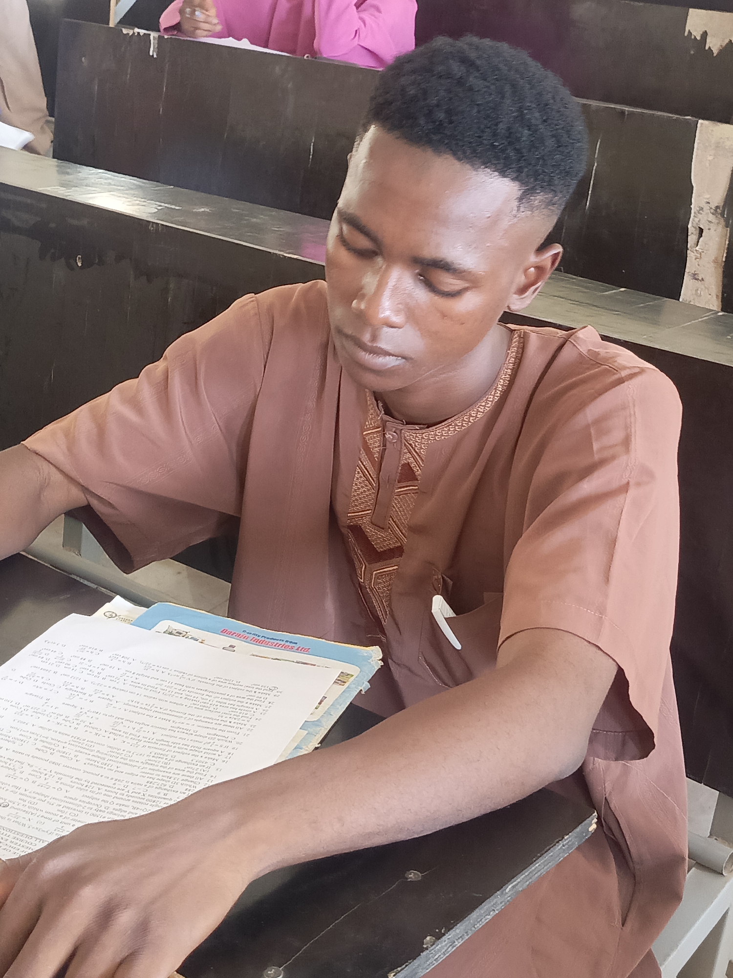 A young man with a short haircut is seated at a desk in a classroom. He is wearing a brown, short-sleeved traditional African shirt with embroidered details around the collar. He appears to be studying, with papers and a textbook in front of him on the desk. The textbook cover is partially visible, displaying "Darain Industries Ltd.". The desk is old and dark, possibly made of wood. He is in a classroom with tiered seating. The location and city of Akwanga, Nigeria are not able to be verified from the image.