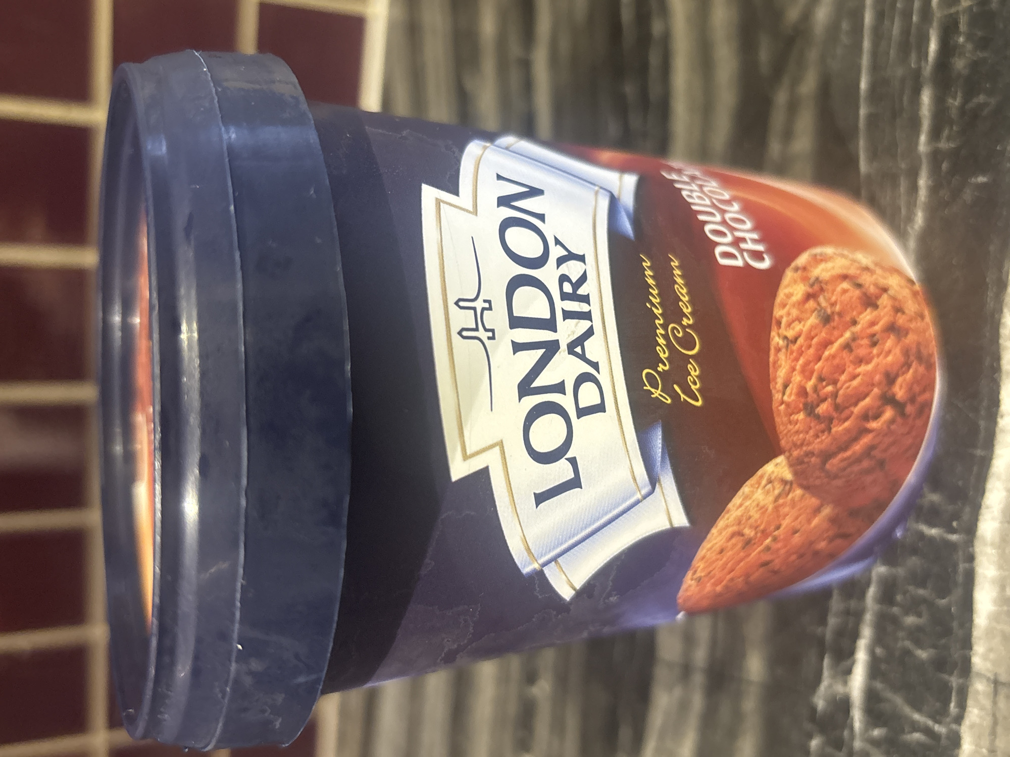 The media file shows a close-up, angled shot of a container of London Dairy premium ice cream. The container is predominantly navy blue with a white label featuring the brand's name, "LONDON DAIRY," in large, dark blue lettering. Above the brand name is a stylized logo resembling a crown. Below the brand name, in smaller gold script, it reads "Premium Ice Cream." To the right of the main label, text in white reads "DOUBLE CHOC," indicating the flavor. The image also shows a realistic depiction of chocolate ice cream, with scoops and swirls, on the right side of the container. The container has a dark blue plastic lid. The background is blurred, but appears to be a tiled surface and a textured, dark surface, possibly a countertop. There are no people visible. The lighting suggests an indoor setting, possibly a kitchen or pantry. The overall impression is a product shot, highlighting the ice cream.