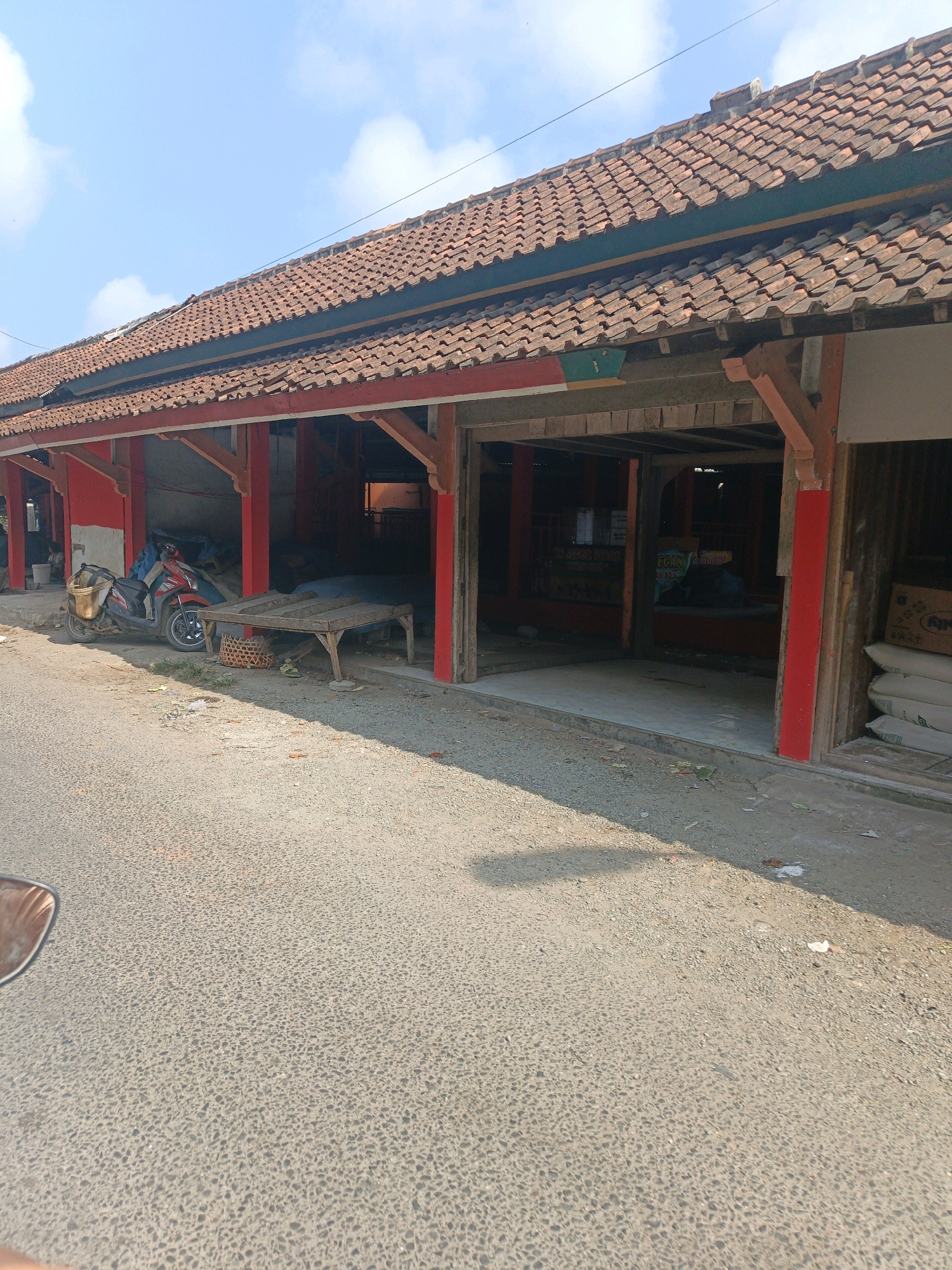 The image captures a street scene in Wetonkulon, Indonesia, on a sunny day. In the foreground, a paved road with a speckled grey surface is visible, with a shadow cast across it from the left. On the right side of the road, a building with a red facade and a tiled roof stands. The building appears to be a row of market stalls or shops with open fronts.
To the left of the building, a red and black motorcycle is parked, equipped with a brown basket on the back. Next to the motorcycle, a wooden table with a woven basket underneath is placed on the ground. The building itself features red-painted support beams and a dark-colored roof. The open stalls reveal dimly lit interiors, some containing stacked white sacks and boxes. The sky above is a clear blue with a few scattered white clouds.