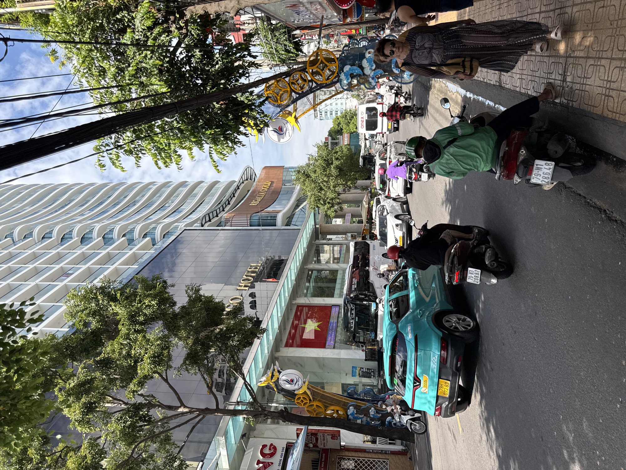 This is a street-level, eye-level, medium shot capturing a busy urban scene in Nha Trang, Vietnam. The image is dominated by a wide, paved street with traffic. In the foreground, a teal-colored electric car, identified as a VinFast VF e34 by its badging and license plate "79H 072.63", is moving down the street. A scooter rider wearing a green jacket and helmet is also visible on a scooter just behind the electric car. Further down the street, other scooters, cars, and a tour bus are present, indicating ongoing traffic flow.
On the right side of the street, a woman with sunglasses and a patterned dress walks along a tiled sidewalk, carrying a bag. Behind her, a section of a building with a glass facade is visible. On the left side of the street, a modern, multi-story building with a distinctive curved architectural design rises high into the sky. This building appears to be a hotel, as indicated by a sign that reads "NG HOTEL". Adjacent to it, another building features a large display of the Vietnamese flag. Trees line the street, providing shade and greenery, and overhead, a dense network of electrical wires crisscrosses the sky. The overall atmosphere suggests a typical day in a bustling Vietnamese city with a mix of modern and traditional elements.