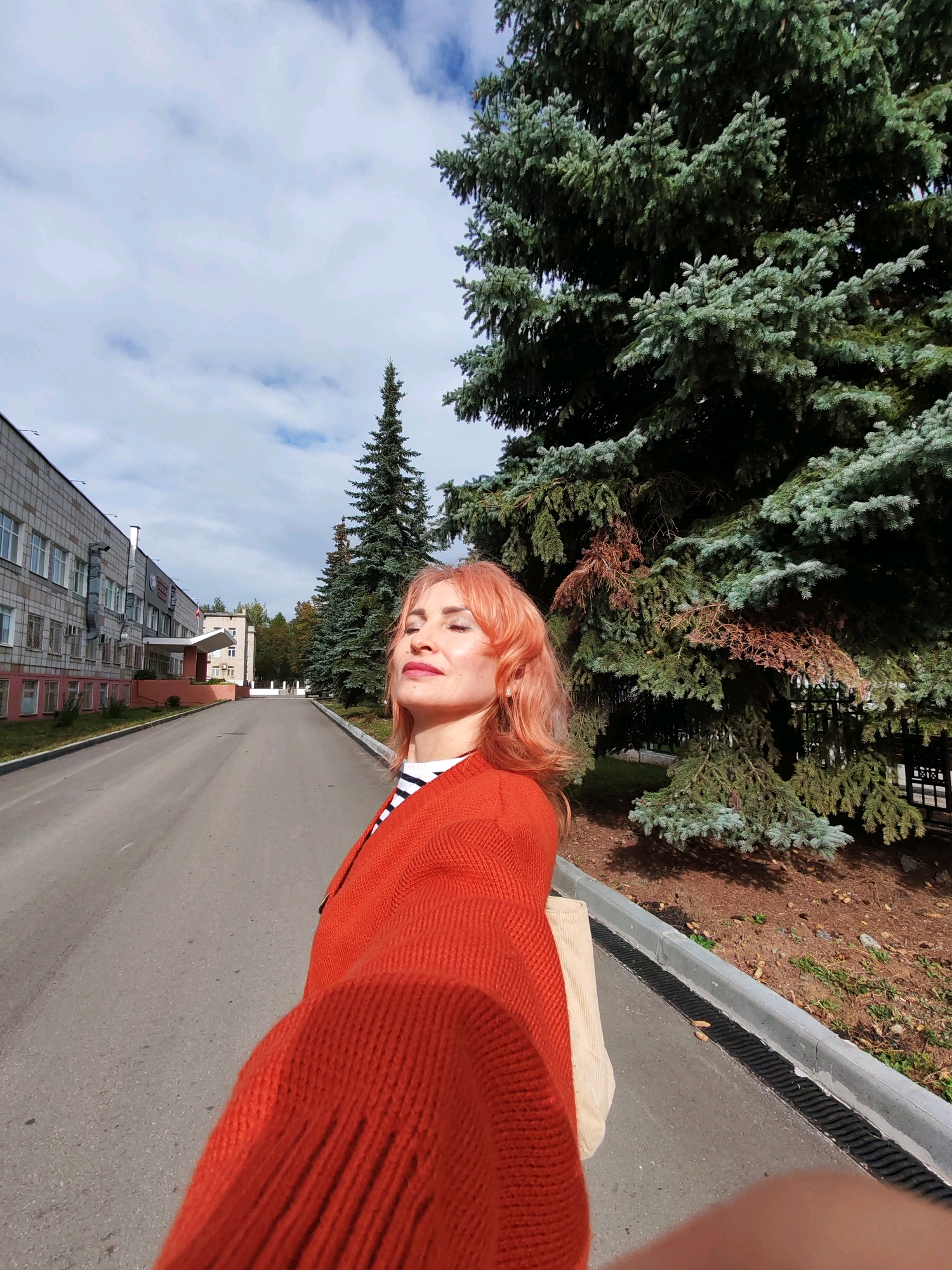 A medium shot, taken from the perspective of someone walking forward, captures a woman with light peach-colored hair and closed eyes, tilted back as if enjoying the sun. She is wearing a bright orange knitted sweater over a striped shirt and carries a beige tote bag. To her left, a long, multi-story building with numerous windows and grey siding extends into the distance, suggesting an institutional or commercial setting. The building's facade is punctuated by red trim at the base and window levels. A paved road, marked with a white line, leads away from the viewer, flanked by a row of tall evergreen trees on the right side of the frame. The trees, with their dense, blue-green foliage, create a natural barrier between the road and what appears to be a manicured area with brown mulch and sparse green ground cover. The sky above is partly cloudy, with patches of blue visible through the white clouds. The overall scene depicts an outdoor environment within an urban or suburban area, likely in Chaikovsky, Russia, given the architectural style and vegetation.