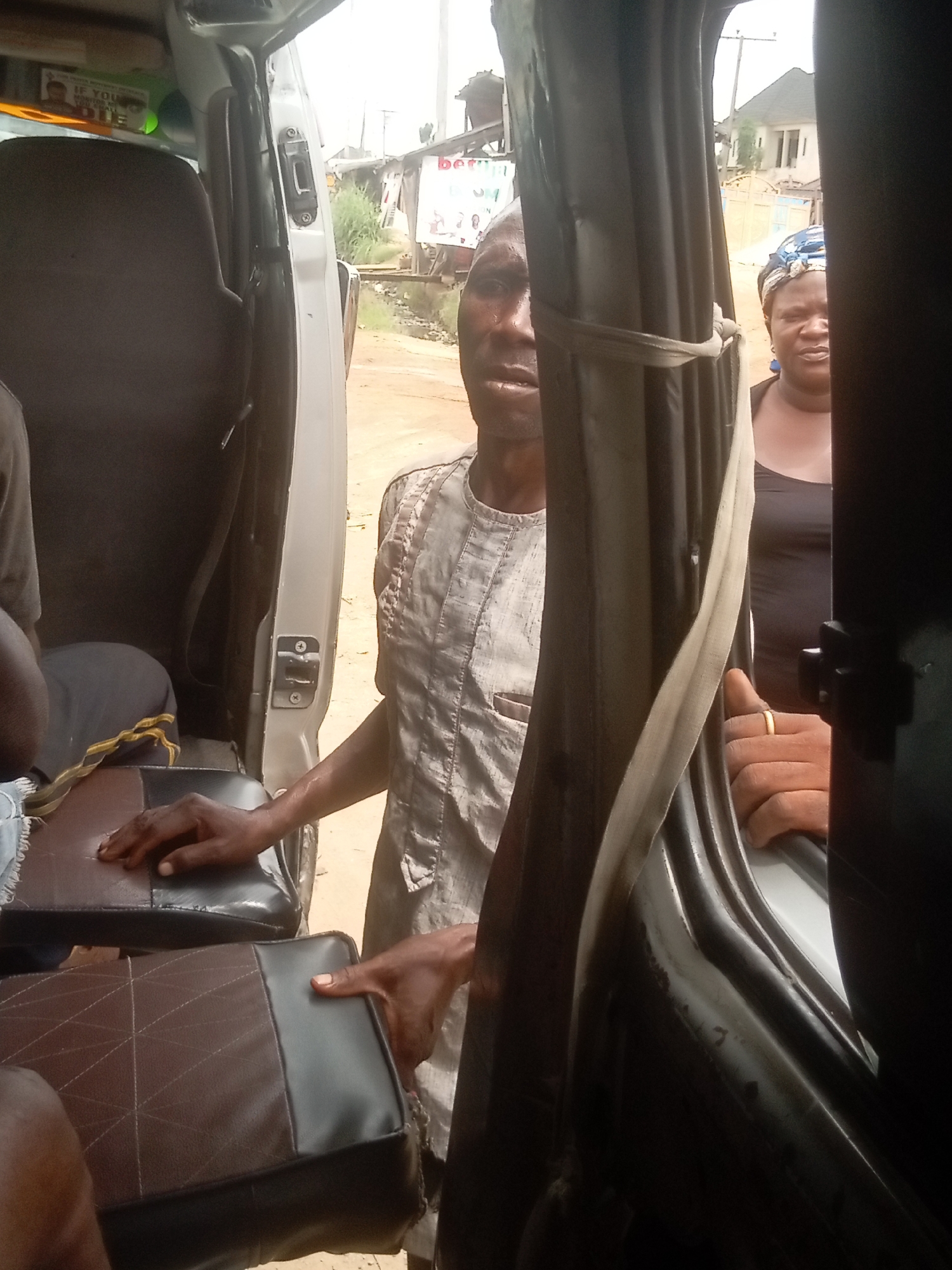 This image depicts a street scene in Orifite, Nigeria, observed from inside a vehicle, likely a commercial passenger van or bus, looking outwards through an open side door during daylight hours.
In the foreground, a man stands outside the vehicle, his torso and head visible, looking towards the camera. He wears a light-colored, possibly traditional, patterned tunic. His left hand rests on the brown and black upholstered seat inside the vehicle, suggesting he is either entering, exiting, or interacting with someone. To his right and slightly behind him, a woman is partially visible, also outside the vehicle, wearing a dark top and a patterned blue and white headscarf, also looking towards the camera. Another person's hand, wearing a gold-colored ring, is visible gripping the interior door frame of the vehicle.
The vehicle's interior features several dark-brown and black seats with a mix of plain and geometric-patterned upholstery. A dark seatbelt is also visible. The background reveals an unpaved dirt road, sparse green vegetation, and distant residential buildings under a bright, possibly overcast, sky.
The activity appears to be a typical moment of public transportation, such as boarding, disembarking, or waiting by the vehicle. The expressions on the visible faces are neutral or contemplative.
Notable details include:
* Inside the vehicle, behind the front seats, a partially visible sign displays fragmented text, including "IF YOU," "MONEY," and "DIE," suggesting a cautionary or religious message.
* In the far background, a blurred sign or billboard shows the letters "bet," possibly indicating a betting service advertisement.
* The setting suggests a common, everyday scene in a semi-urban or rural area of Nigeria.