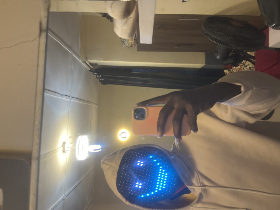 A person in a hooded sweatshirt and an LED smiley-face mask takes a selfie in a mirror. The mirror reflects a room with a dresser, a fan, and curtains. The hand holding the phone is dark-skinned. The mask's display features a bright blue smiling face. A light fixture is visible on the left side of the mirror.