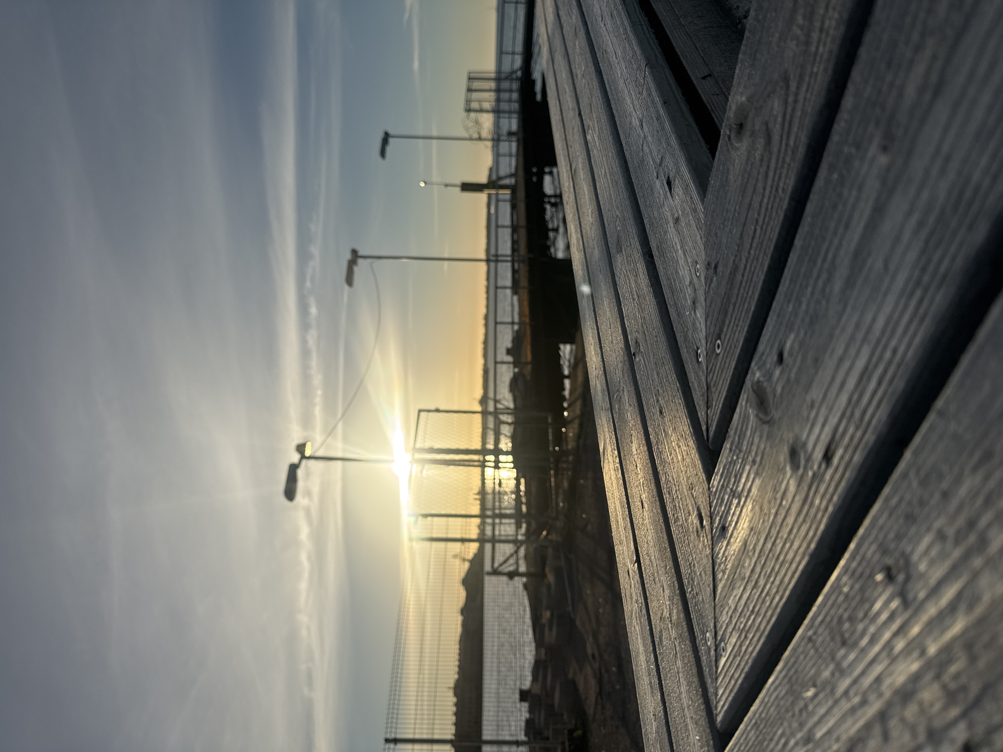 The image depicts a low-angle, close-up view of wooden planks in the foreground, suggesting a pier or boardwalk. The planks are weathered and show a visible grain pattern, with some screws securing them. Beyond the wooden structure, a body of water is visible, likely the sea, with a distant shoreline featuring industrial structures or buildings. Two lampposts are present, one closer and one further away, with lights illuminating the sky. The sun is low on the horizon, casting a warm glow and creating lens flare effects. The sky is a blend of blue and muted orange, with wispy clouds. The scene appears to be set in Tallinn, Estonia, possibly at a harbor or waterfront area.