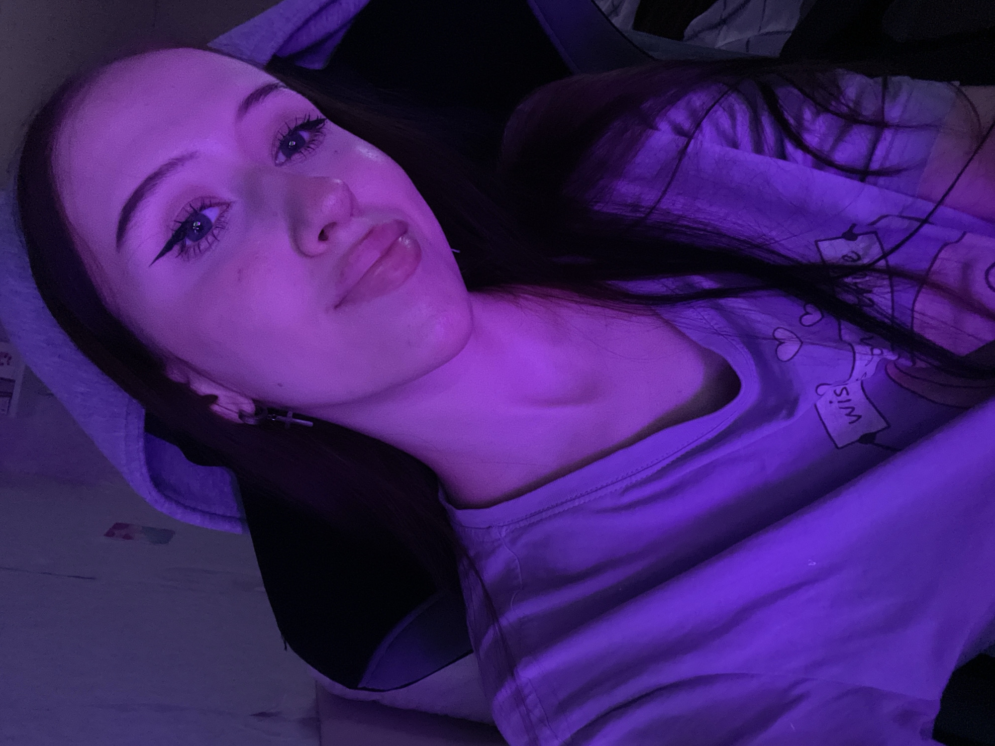 A young woman with long dark hair and dark eye makeup is pictured from the chest up, looking at the camera. She is wearing a light-colored t-shirt with a graphic of hearts and text. The scene is illuminated by purple lighting, casting a glow on her skin and surroundings. She appears to be in an indoor setting, possibly a room with a chair visible behind her. The image does not provide any specific information about Votkinsk, Russia, or any other location.