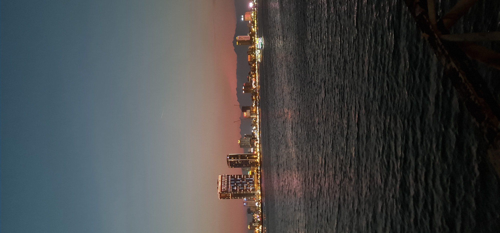 This image presents a panoramic view of a coastal city at twilight or early night, looking across a body of water. The scene is dominated by a clear sky, a vibrant cityscape, and the reflective surface of the water.
The upper portion of the image features a gradient sky, transitioning from a deep blue-grey at the top to a warm, reddish-orange glow along the horizon, characteristic of dusk. A faint, thin white line, possibly an airplane contrail or a distant meteor, is visible in the upper-left area of the sky.
Along the horizon, a bustling cityscape stretches across the frame. Numerous multi-story buildings, appearing as hotels or residential towers, are brightly illuminated with yellow and white lights, casting a warm glow and creating intricate patterns on their facades. Behind the illuminated city, the dark silhouettes of mountains are visible, subtly touched by the reddish hues of the twilight sky.
The lower and right-hand side of the image is dominated by the expansive surface of a body of water, likely a sea or a large river. The water exhibits ripples and small waves, reflecting the city lights as shimmering streaks. On the extreme right edge of the frame, a dark, indistinct structure, possibly a railing or part of a pier, is partially visible. The overall scene evokes a sense of urban tranquility as the city comes alive with lights after dusk, consistent with a vibrant coastal city like Da Nang, Vietnam. No discernible text is visible in the image.