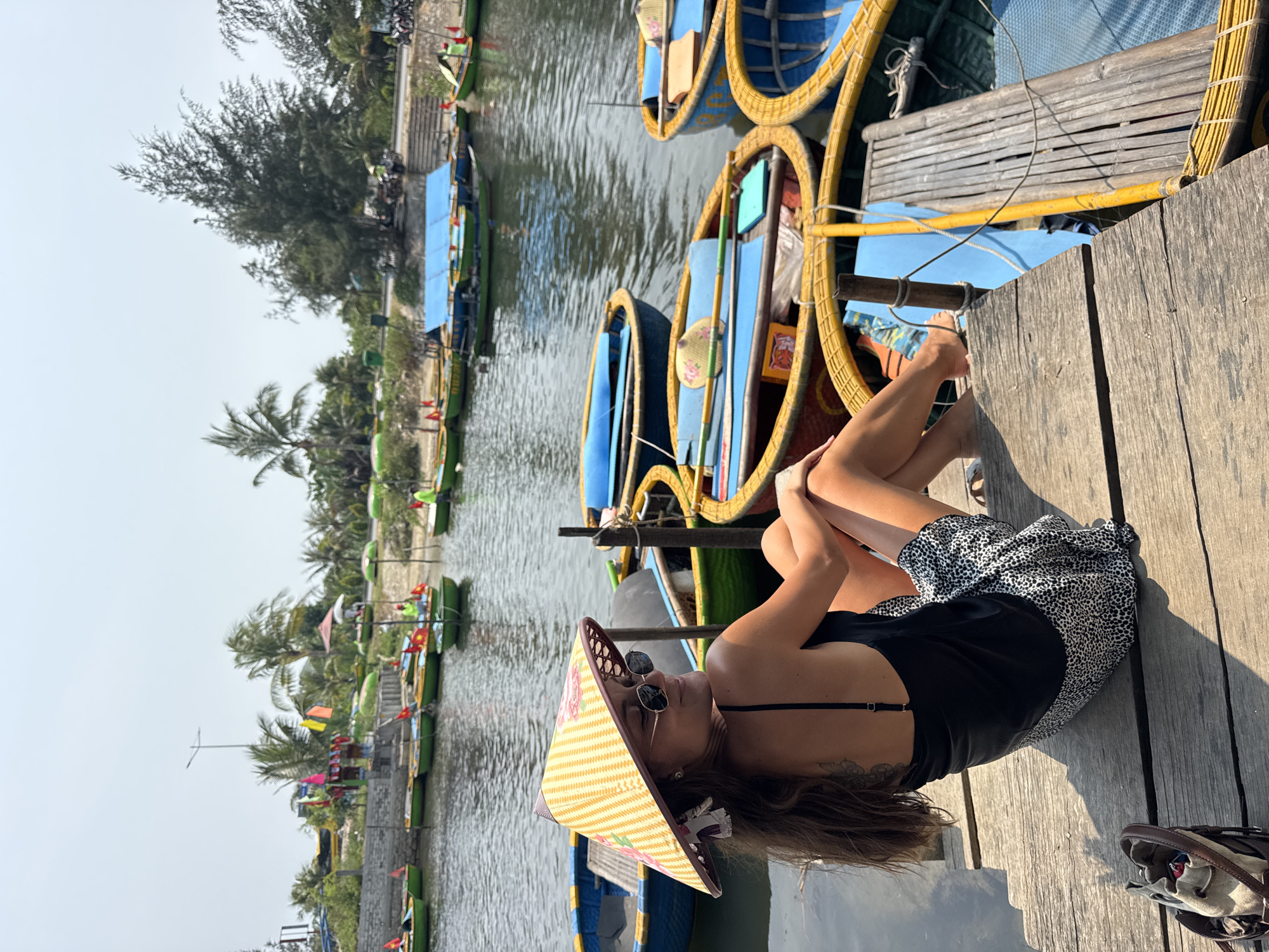 The image portrays a bright, sunny outdoor scene in Đà Nẵng, Vietnam, featuring a woman relaxing on a wooden dock beside a calm river.
The main subject is a woman, casually lying on her side on the weathered wooden planks of the dock. She is wearing a black tank top, a black-and-white leopard print skirt, and sunglasses. Her long brown hair flows around her, and she sports a distinctive wide-brimmed conical hat with yellow stripes and pink floral patterns. A tattoo is visible on her left side/back. A small, light-colored bag with brown straps is placed next to her on the dock.
Alongside the dock and in the river, numerous traditional Vietnamese basket boats, known as Thung Chai, are moored. These boats are brightly colored, primarily yellow and blue, and some have small, decorative fans attached. Further down the river, several green, rectangular boats with blue coverings are also visible. The river water is calm, reflecting the daylight.
The far bank of the river is lush with vegetation, including several tall palm trees and other foliage, typical of a tropical environment. A pathway or road appears to run along the far bank. The bright sunlight indicates a clear, pleasant day, casting clear shadows on the dock and boats. The overall scene conveys a sense of leisure and tranquility in a picturesque waterfront setting. No discernible text is visible in the image.