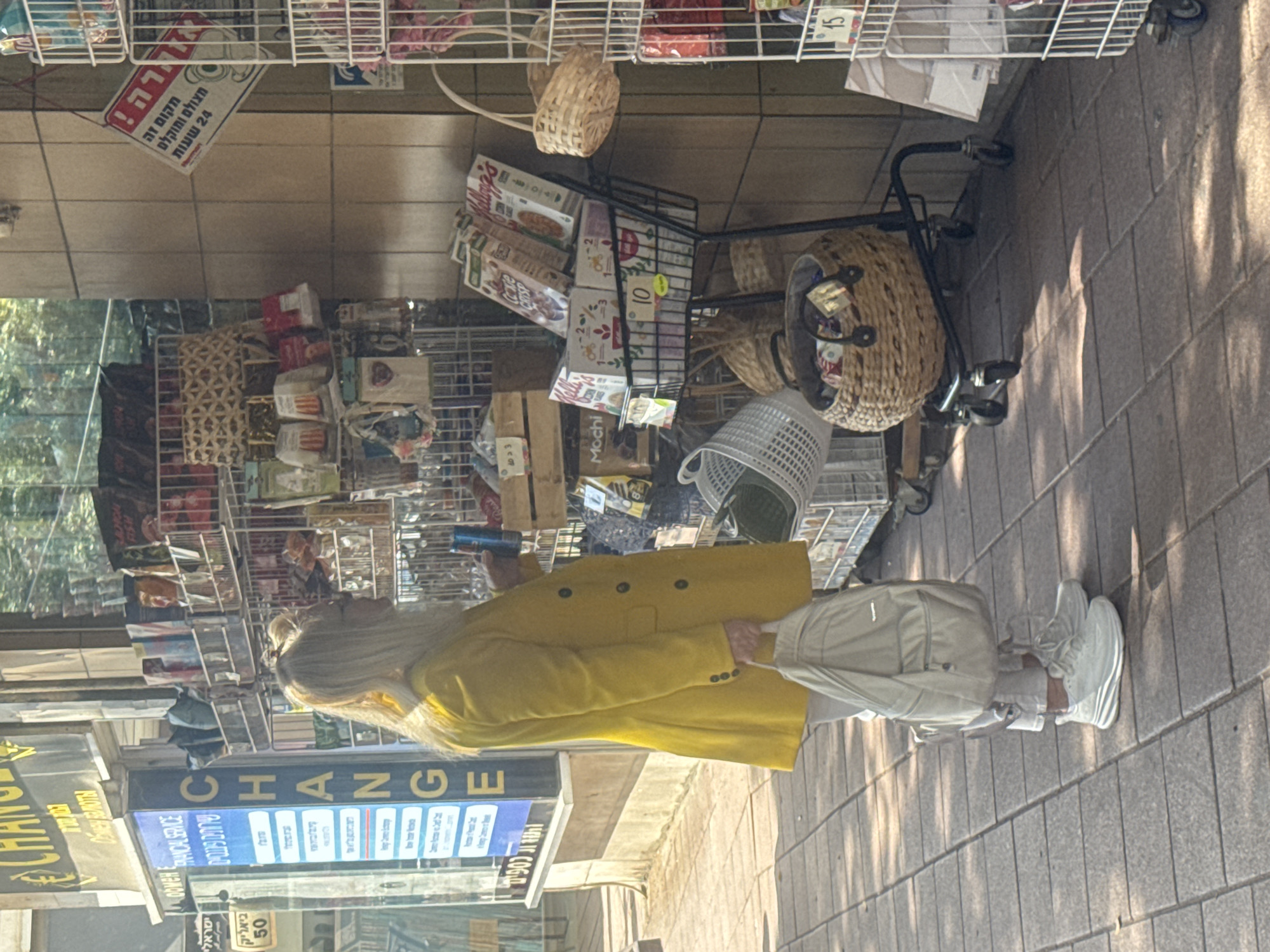 Here is a detailed description of the image:
**General Description:** The image shows a street scene with a person walking past a shop. The scene appears to be set outdoors, possibly in a commercial district.
**People:** One person is clearly visible in the image. They are wearing a yellow coat, white pants, and white sneakers. They are walking on the sidewalk.
**Objects:** The image contains several objects including a shopping cart filled with various items, shelves with merchandise, signage, and an awning.
**Actions:** The person is walking past the shop. There is a sign that says "CHANGE".
**Interactions:** The person is interacting with the environment, specifically walking past the shops on the street.
**Environment:** The environment is an outdoor street setting, with a shop storefront and sidewalk. The image indicates a sunny day with shadows cast on the ground.
**Location Details:** Based on the signage in Hebrew ( "ישראלי ביאליק 50") and the OCR-detected signs, the image appears to be taken in Ramat Gan, Israel. The text seems to reference Bialik street. The sign also states: "This place is photographed and recorded 24 hours".