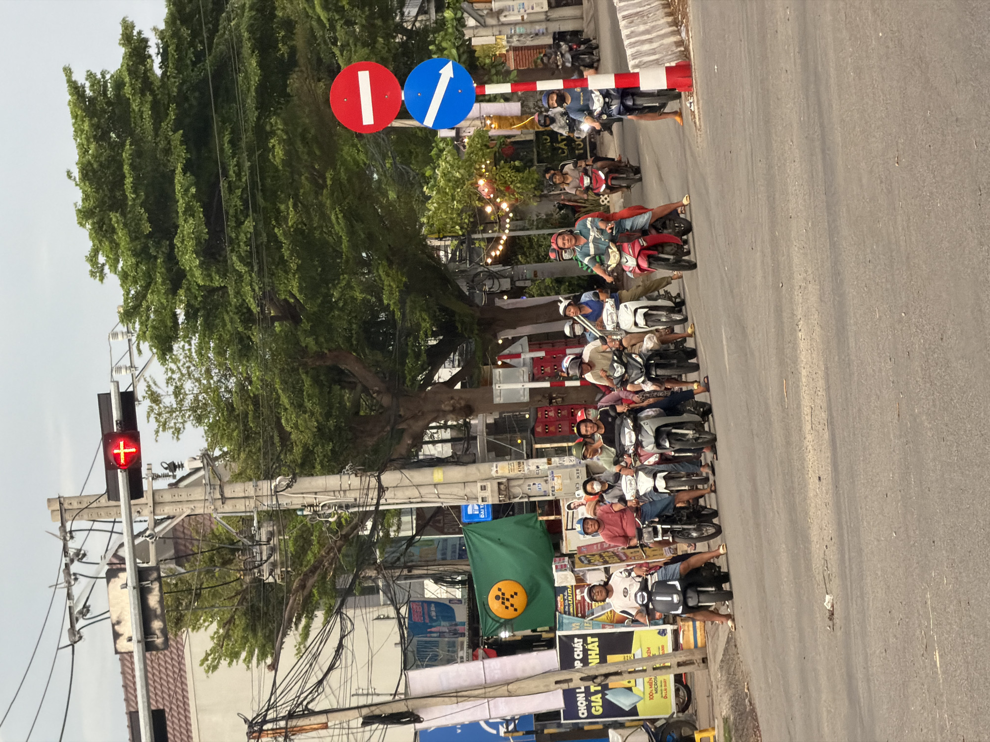 The vertically oriented image captures a bustling street scene in Dong Hai Ward, Vietnam, likely an intersection or a point where traffic is halted. A large group of people, primarily on scooters and motorcycles, are seen waiting in lines, facing towards the right side of the frame. Most individuals are wearing helmets, suggesting they are adhering to local traffic regulations.
In the mid-ground, a red traffic light displays a red "plus" or "cross" symbol, indicating that traffic should stop. Next to it, two circular traffic signs are prominent: a red sign with a white horizontal bar signifying "no entry," and a blue sign with a white upward arrow indicating "straight ahead." A red and white striped barrier runs across the lane, further reinforcing the stop.
The street is lined with lush green trees, and some decorative string lights are visible among the foliage, suggesting it could be late afternoon or early evening. Overhead, a complex network of power lines and utility poles crisscrosses the scene. Buildings with typical urban architecture can be seen along the roadside. A green flag with a yellow circular logo featuring an orange, stylized 'W' or 'M' hangs prominently near the center.
Advertising banners and posters are visible on the left side of the street, displaying Vietnamese text such as "CHỌN L OP CHẤT," "GIÁ T NHẤT," "100% MIỀN," "MICROS," "ON LÃI SUẤT," "KEM," "UUDA," "DAITAB," and "CẮT." The overall atmosphere appears calm, with people patiently waiting for the traffic to proceed.