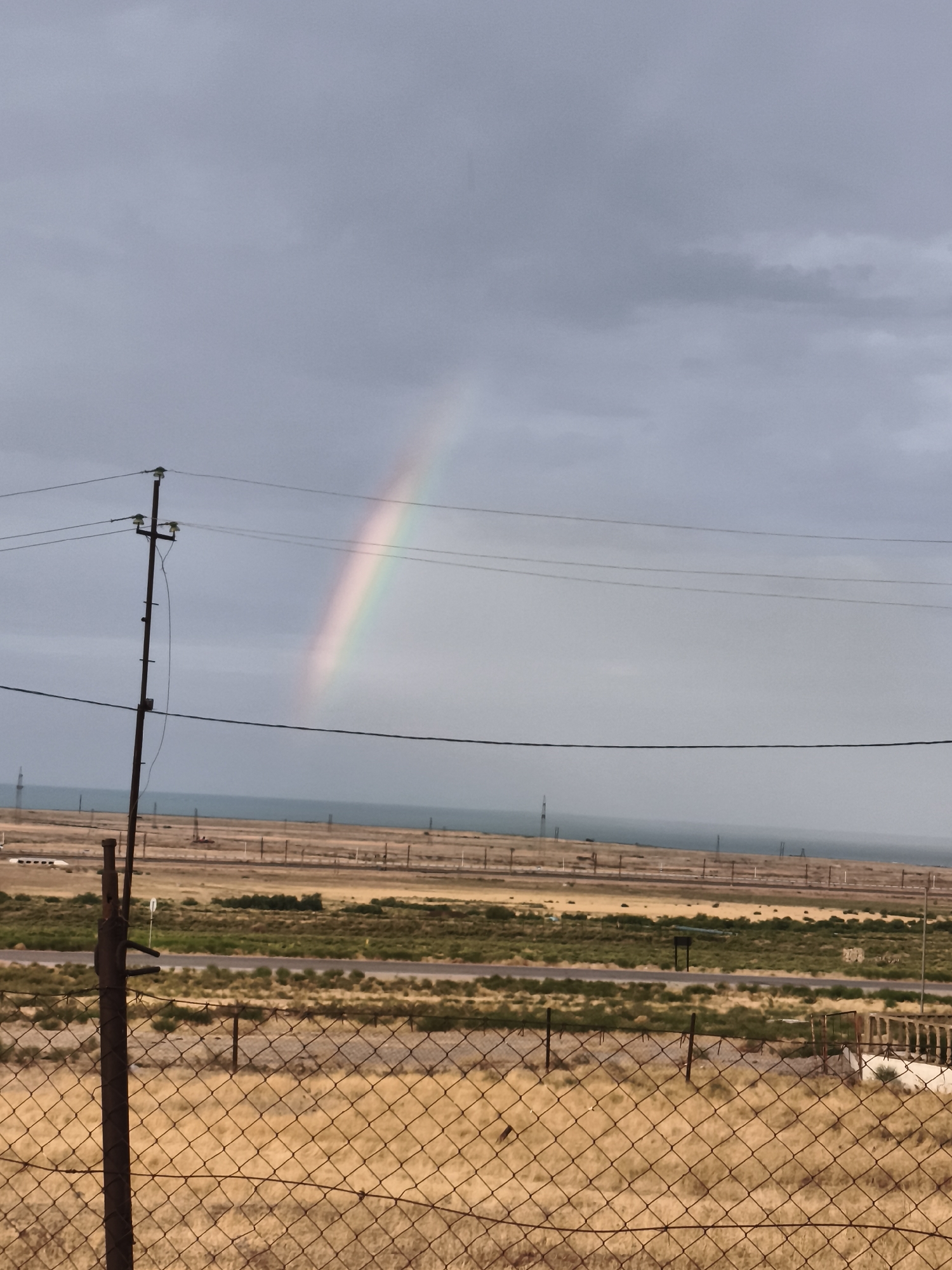 The image depicts a landscape with a rainbow arching across a cloudy sky. In the foreground, a rusty metal fence with diamond-shaped mesh encloses dry, yellowed grass. Behind the fence, a power pole with insulators and wires stands prominently on the left. Beyond that, a flat, arid landscape stretches out with sparse vegetation, dirt roads, and distant industrial structures or power lines. The horizon is marked by a body of water, possibly the Caspian Sea, under a gray, overcast sky. The overall impression is one of a somewhat desolate, rural or industrial outskirts, with the rainbow offering a transient splash of color. The scene is devoid of people or discernible activity.
