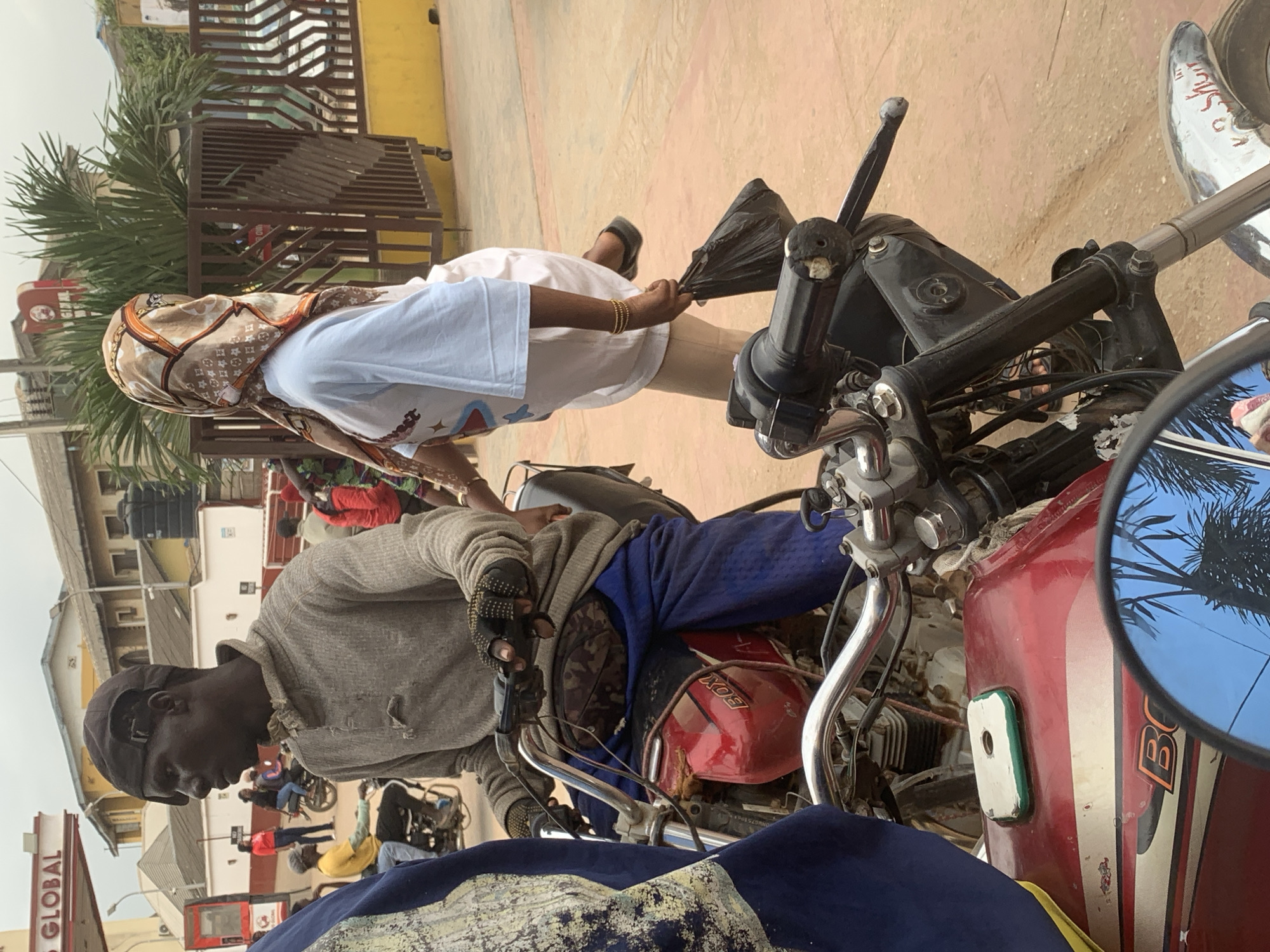 The image depicts a street scene in Shagamu, Nigeria, likely near a gas station with a "Global" sign visible. In the foreground, two motorcycles are parked side-by-side. The motorcycle on the left is partially obscured, showing a red fuel tank with a "BOX" logo. The rider of this motorcycle is wearing a grey long-sleeved shirt, blue pants, and a dark cap. They are also wearing camouflage-patterned fanny pack around their waist and black fingerless gloves. To the right of the first motorcycle rider, a person wearing a white t-shirt and a patterned headscarf is walking, carrying a black plastic bag in their right hand. They are wearing a bracelet on their left wrist. In the background, other individuals and motorcycles are visible, along with buildings and a yellow fence. The ground is a dusty, reddish-brown color.
