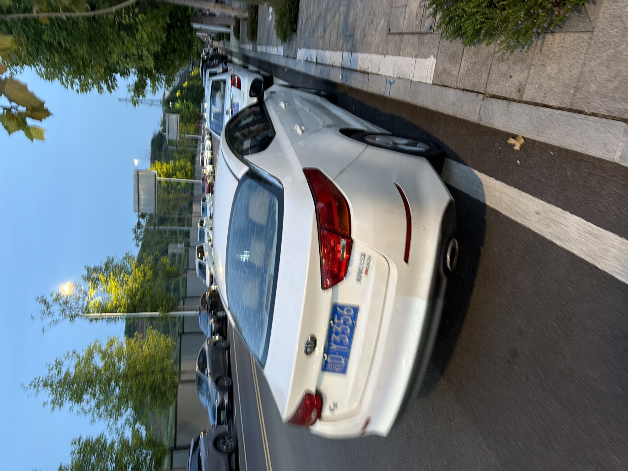 The image depicts a street scene at dusk in Jiangjin District, Chongqing, China. A white Kia sedan is the prominent subject, parked on the right side of a road. The car's license plate is visible and reads "渝D Y3356". Several other cars are parked along the street and on the road. Trees line the sidewalk, and a street lamp is illuminated in the background. The sky is a clear blue, suggesting the end of daylight. The overall impression is of a quiet, suburban or semi-urban street at the end of the day.