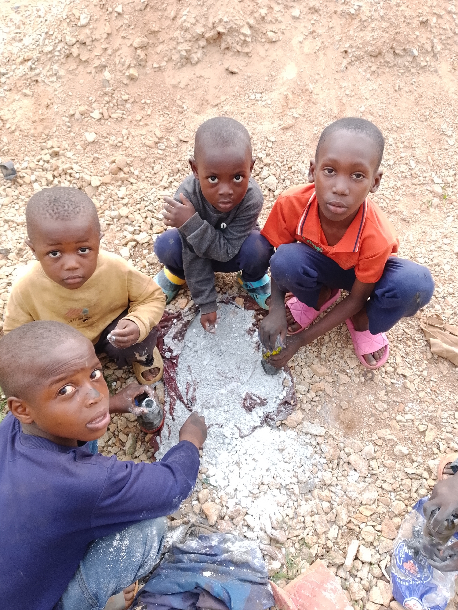 In this medium shot, four young children are crouched on a rough, gravelly ground. The children, with dark skin and short hair, appear to be playing or working with a mixture of grey powder and some red material spread on a piece of dark fabric.
The child on the far left, wearing a yellow long-sleeved shirt and blue jeans, looks directly at the camera with an inquisitive expression. Next to him, a child in a grey hooded sweatshirt and blue pants is also looking at the camera, holding a small object in their hands.
To the right, a child in an orange collared shirt and blue pants is actively engaged with the mixture, using a stick or tool to interact with it. Their pink sandals are visible. The fourth child, closest to the viewer, is wearing a dark blue long-sleeved shirt and blue jeans, and is also looking towards the camera, holding a small grey object.
The ground is covered in small stones and dirt, suggesting an outdoor, possibly rural or undeveloped setting. The lighting suggests it is daytime, with no specific indications of weather conditions. The expressions on the children's faces are neutral, perhaps a mix of concentration and curiosity. There is no visible text in the image. The overall scene depicts children engaged in an activity that could be a game or a rudimentary form of work involving mixing materials.