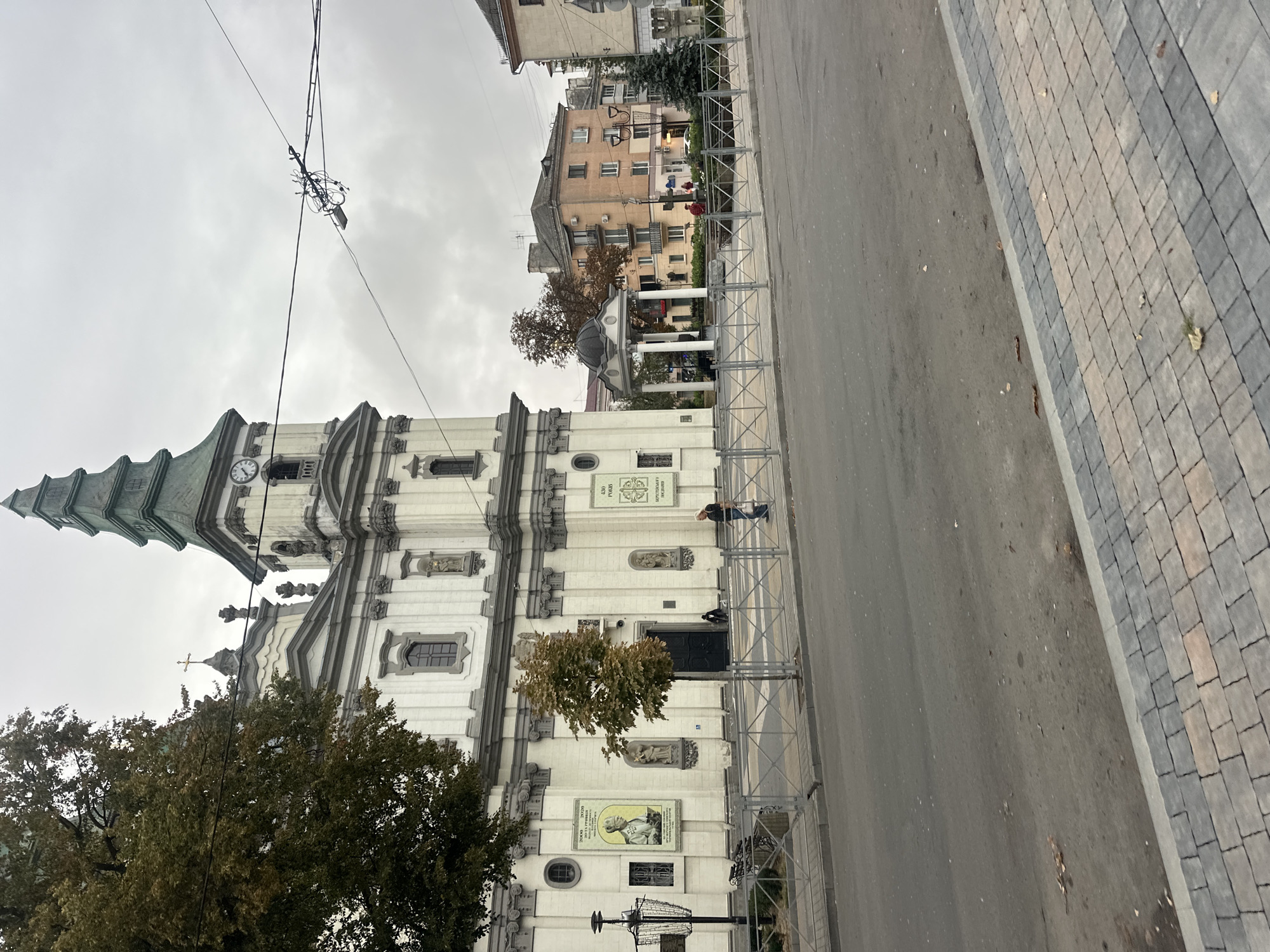 The image shows a street view of the Ternopil Regional Philharmonic, formerly the Church of the Immaculate Conception of the Blessed Virgin Mary, located in Ternopil, Ukraine. A large white church with a green spire and clock tower dominates the left side of the frame. A tree with autumn foliage stands in front of the church. A metal fence separates the sidewalk from the street. A person is walking on the sidewalk to the right of the tree. In the background, several buildings, including apartment blocks, are visible. The sky is overcast.