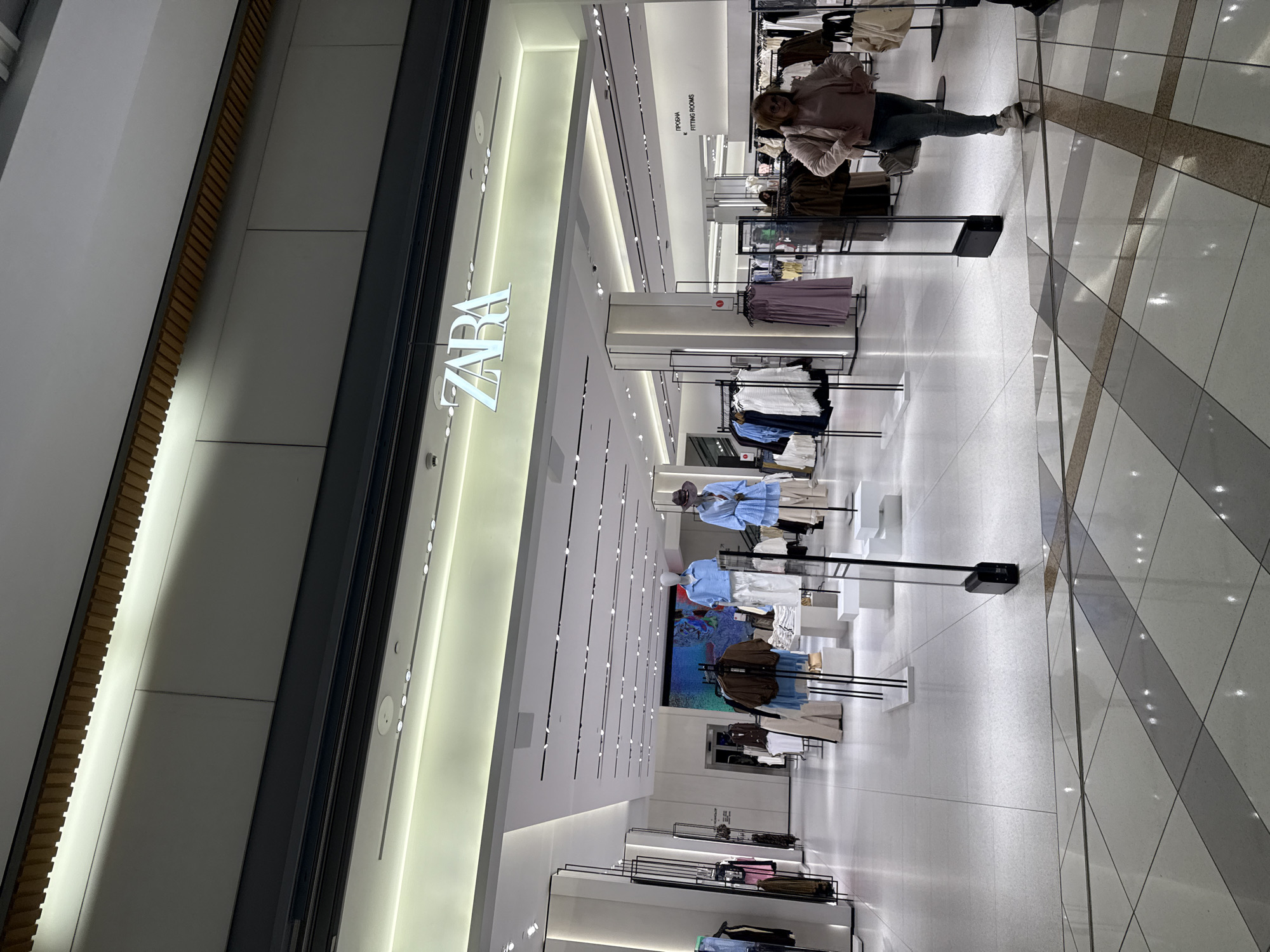 The image displays the brightly lit interior of a Zara clothing store. The prominent "ZARA" logo, illuminated in white, is visible on a large vertical panel that forms part of the store's entrance or main display wall on the left side of the frame. The store boasts a modern and spacious design with white walls, ceilings, and light-colored flooring. Numerous recessed spotlights and linear lighting fixtures adorn the ceiling, ensuring excellent illumination throughout the space.
In the mid-ground, a woman, likely a customer, stands towards the right side of the store. She is dressed in a light jacket and dark trousers, appearing to be observing the merchandise or waiting. Several mannequins are strategically placed around the store, showcasing various outfits, including light blue blouses, brown jackets, and neutral-toned trousers. Numerous clothing racks filled with garments are arranged across the store floor.
A white sign, hanging from the ceiling above the merchandise, clearly indicates "ПРОБНИ / FITTING ROOMS" in both Cyrillic and English. The floor is composed of light grey and off-white tiles, some arranged in a diagonal pattern, reflecting the overhead lights and contributing to the clean aesthetic. The scene captures a typical shopping environment within a contemporary retail establishment in Varna, Bulgaria.