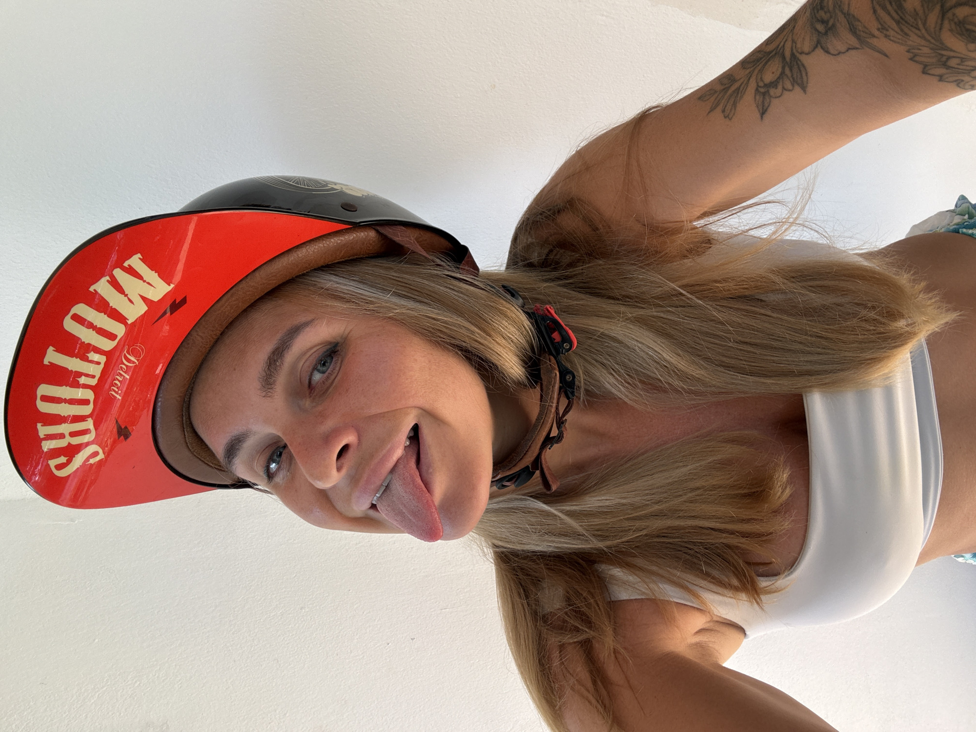 A blonde woman wearing a white tank top and a red and black motorcycle helmet with "Detroit Motors" written on it, sticks her tongue out. Her hair is long and blonde, and she has a tattoo on her arm. She is against a white wall. The scene appears to be in a city, possibly Ko Samui District, Thailand.