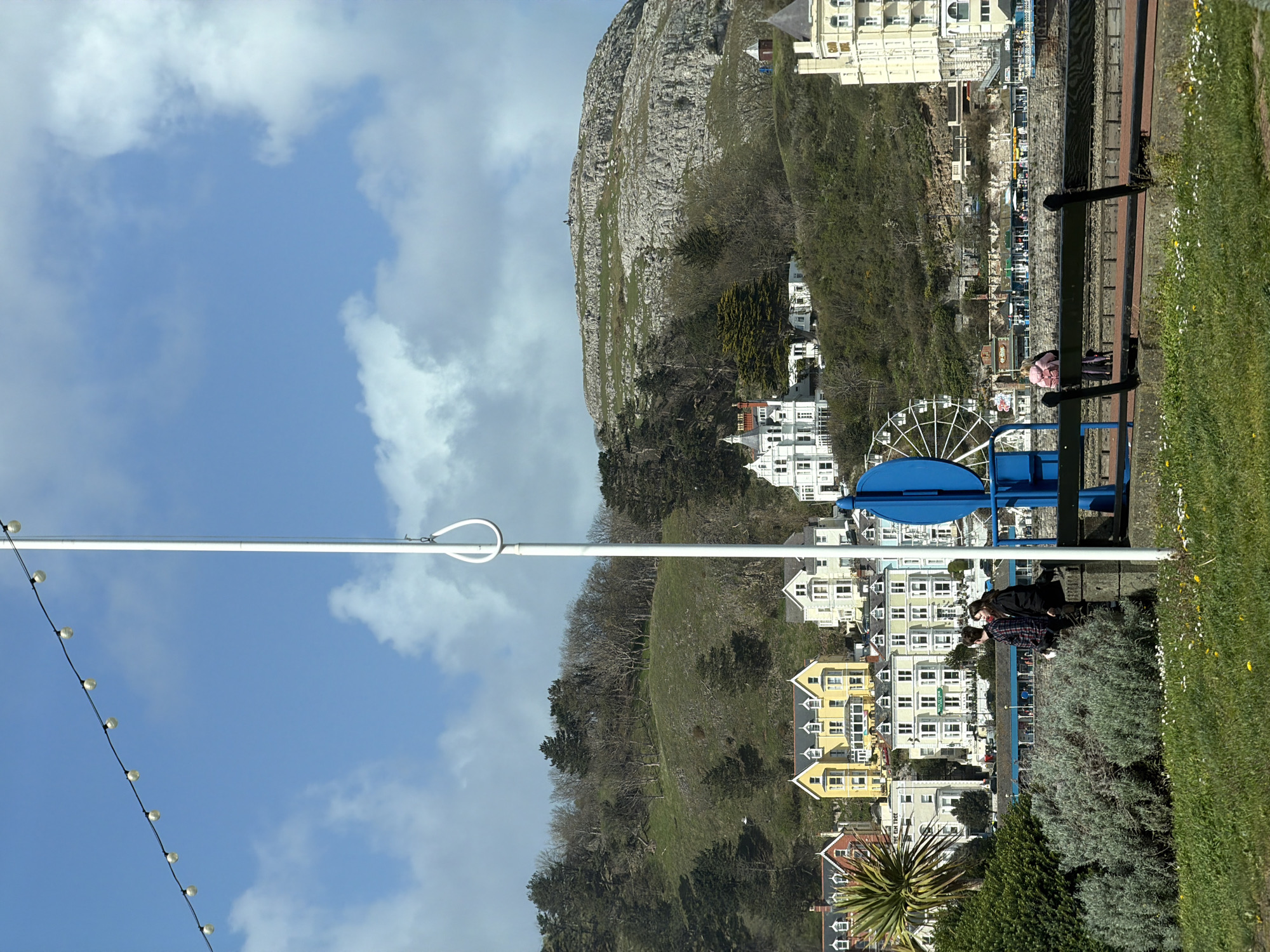 Captured in Llandudno, United Kingdom, the scene showcases a blend of natural and urban elements under a partially cloudy sky. A rocky, green-covered mountain forms a significant backdrop. At its base, a cluster of white and yellow residential buildings stands, with a variety of architectural styles evident. In the foreground, on a patch of green grass and vegetation, sit a group of young people. Some metal railings and a piece of blue fairground or playground equipment are visible. A white pole with lights extends into the blue sky, adding a linear element to the composition. The overall perspective is slightly rotated, altering the usual orientation of the landscape.