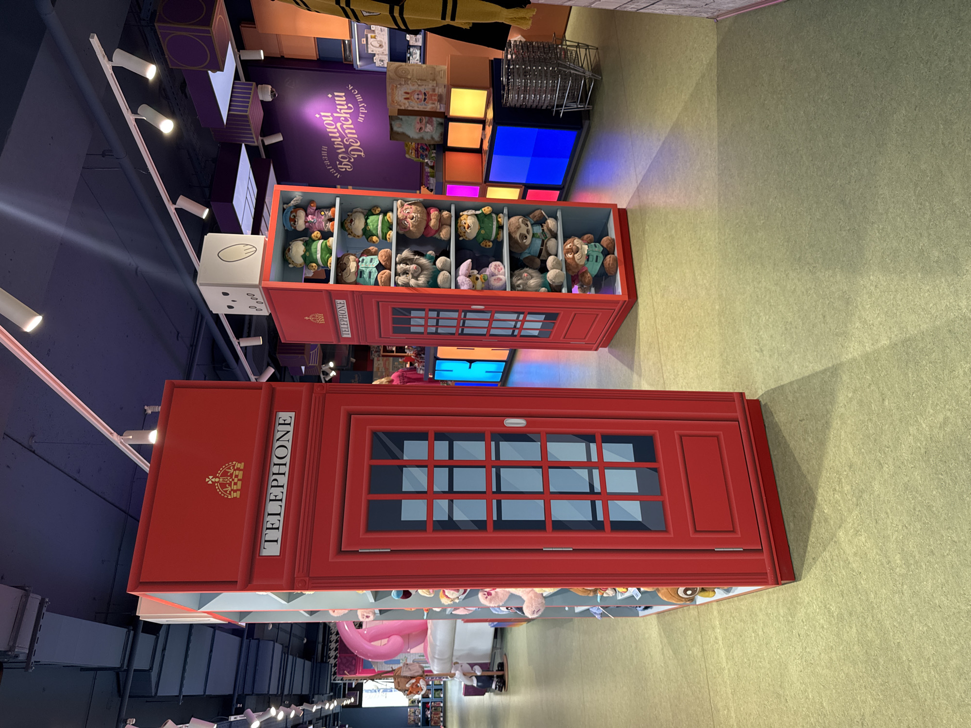 The image shows an indoor setting, possibly a shop or an exhibition. The primary focus is a large, red, British-style telephone box, which appears to be a display or a piece of furniture. Inside the upper section of the telephone box, several stuffed animals are visible. Below the telephone box, there is a shelf filled with more stuffed animals and some other items.
In the background, there are shelves displaying various items, including what appear to be toys or decorative objects. There is also a purple wall with Cyrillic text, which reads "Магазин Большой Детский" (Magazin Bol'shoy Detskiy), meaning "Big Children's Store." Colorful illuminated panels are also visible behind the telephone box. The overall scene suggests a bright and playful environment, likely a children's store. The lighting appears to be artificial, typical of an indoor commercial space. The image is taken from a slightly elevated perspective, looking down and across the scene. The objects are well-lit, and there are no visible people. The flooring is a light-colored carpet or similar material. The telephone box itself is a prominent object, designed to resemble a classic red K6 telephone kiosk, complete with the "TELEPHONE" signage and a crown emblem.
