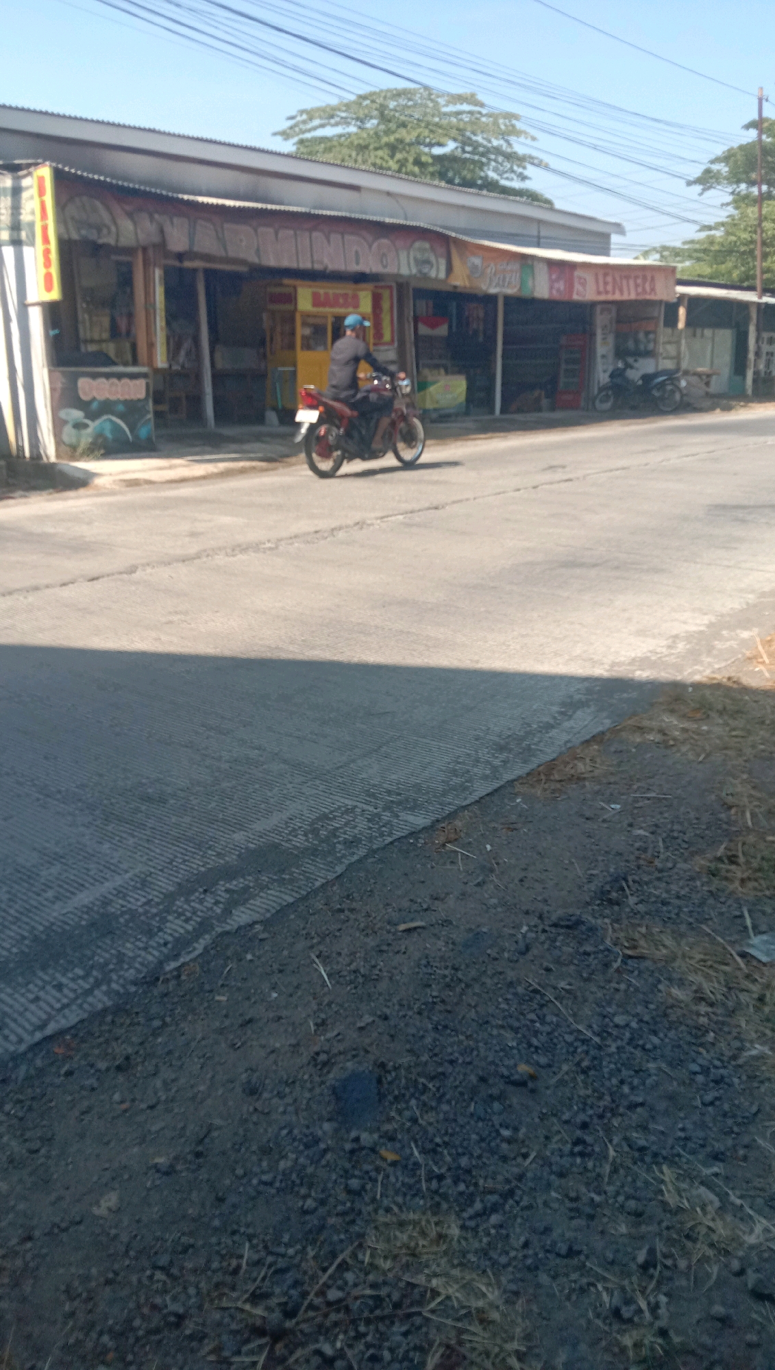 A man wearing a blue cap and dark clothing is riding a red motorcycle on a paved road. The man is facing to the left of the frame. To the right of the road is a row of buildings with awnings, several of which have signage. One sign reads "LENFERA". A motorcycle is parked in front of one of the buildings. To the left of the road is a sidewalk with a striped pattern. In the background, trees and power lines are visible under a clear blue sky. The foreground of the image shows the road surface and some gravel.