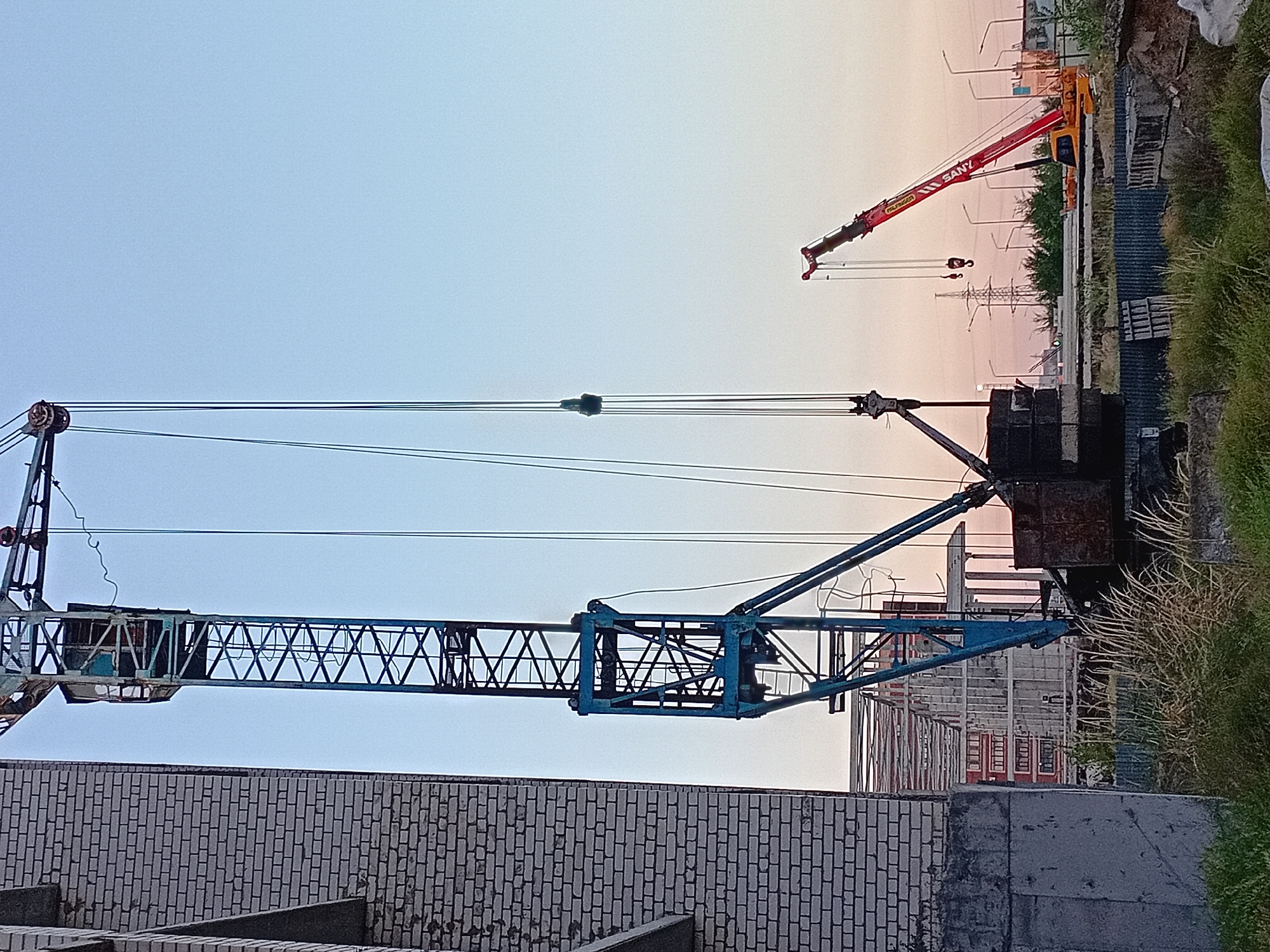 The image shows a construction site in Semey, Kazakhstan. A large blue tower crane stands on the left, with its jib extending upwards and to the right. Below the jib, a dark, rectangular container is suspended by cables. To the right, a smaller red and yellow mobile crane, identified by the "SANY" logo on its boom, is positioned with its boom raised. In the background, several streetlights and power lines are visible against a pale blue and pink sky, suggesting it is either dawn or dusk. A partially constructed building with a brick facade is seen on the far left. The ground is covered with dry grass, debris, and some concrete blocks.