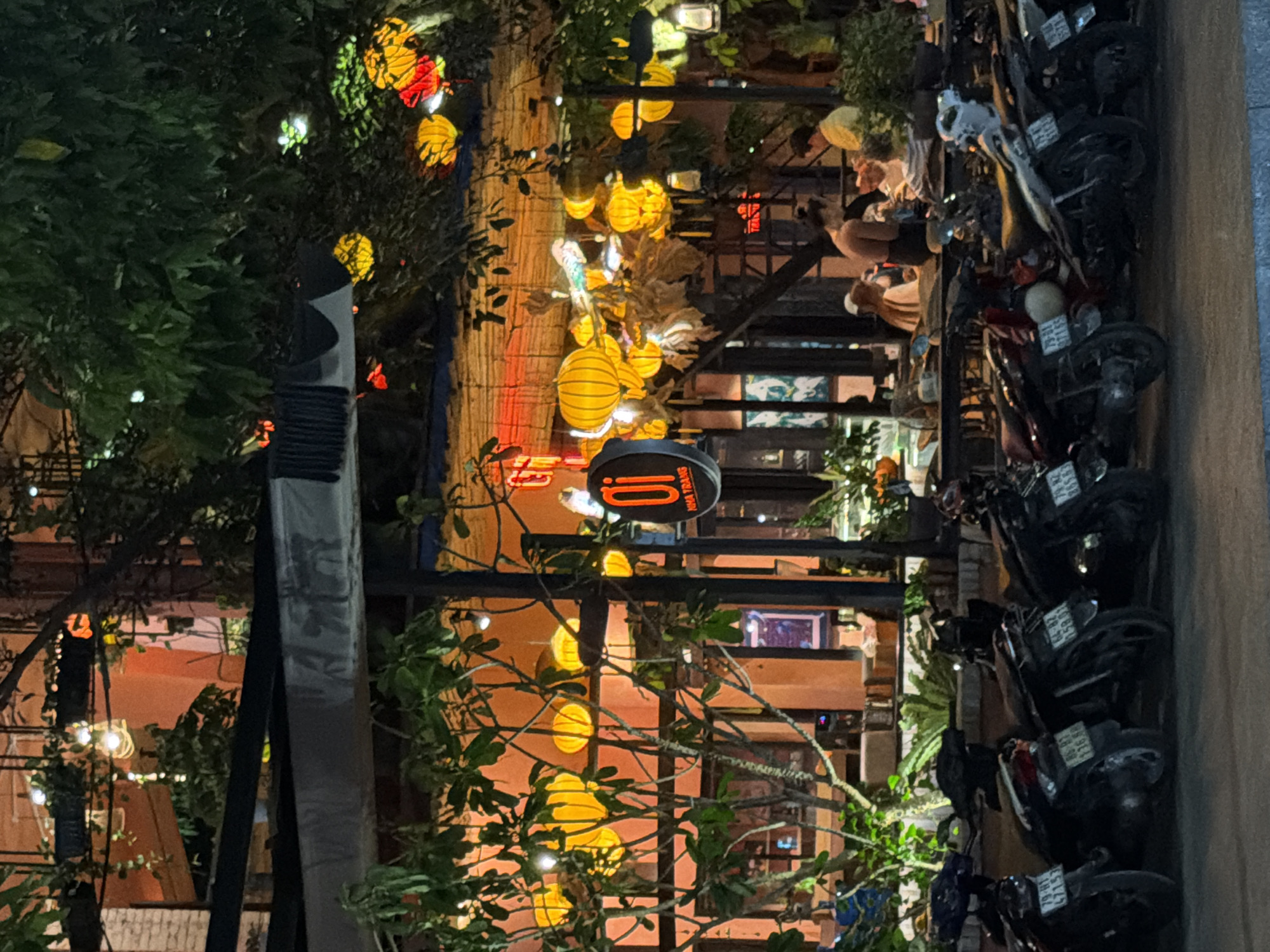 The image depicts an outdoor seating area of a restaurant or cafe in Nha Trang, Vietnam, at night. The establishment is decorated with numerous bright yellow lanterns and some red ones, creating a warm and festive atmosphere. Lush green foliage is visible, interspersed with the decorative lighting. In the foreground, several parked motorcycles are lined up. Behind them, through the branches and lighting, the interior of the establishment can be partially seen, with patrons seated at tables. Some individuals are visible, appearing to be engaged in conversation or dining. The overall impression is of a lively nighttime scene in a Vietnamese city.