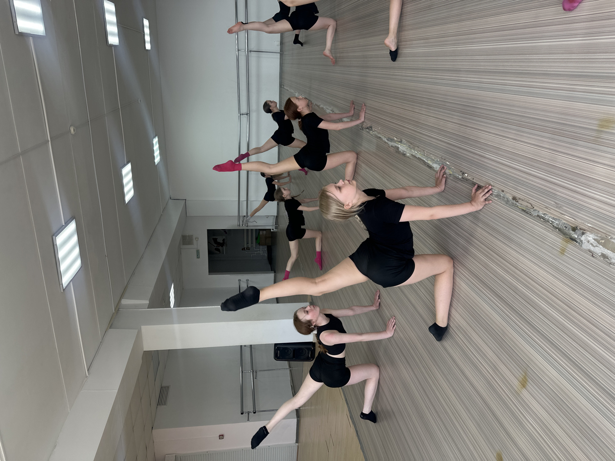 The image shows a dance studio with several young women in black dance attire performing a stretching exercise. The studio has a light-colored, wood-patterned floor and white walls. A row of fluorescent lights is visible on the ceiling. The women are positioned in a line along the wall, touching the wall with their hands for balance while performing a split or leg extension. A speaker is visible against the wall in the background. Based on the provided context, the location is likely Votkinsk, Russia, but there are no specific details in the image to confirm this. The activity appears to be a dance class or rehearsal.