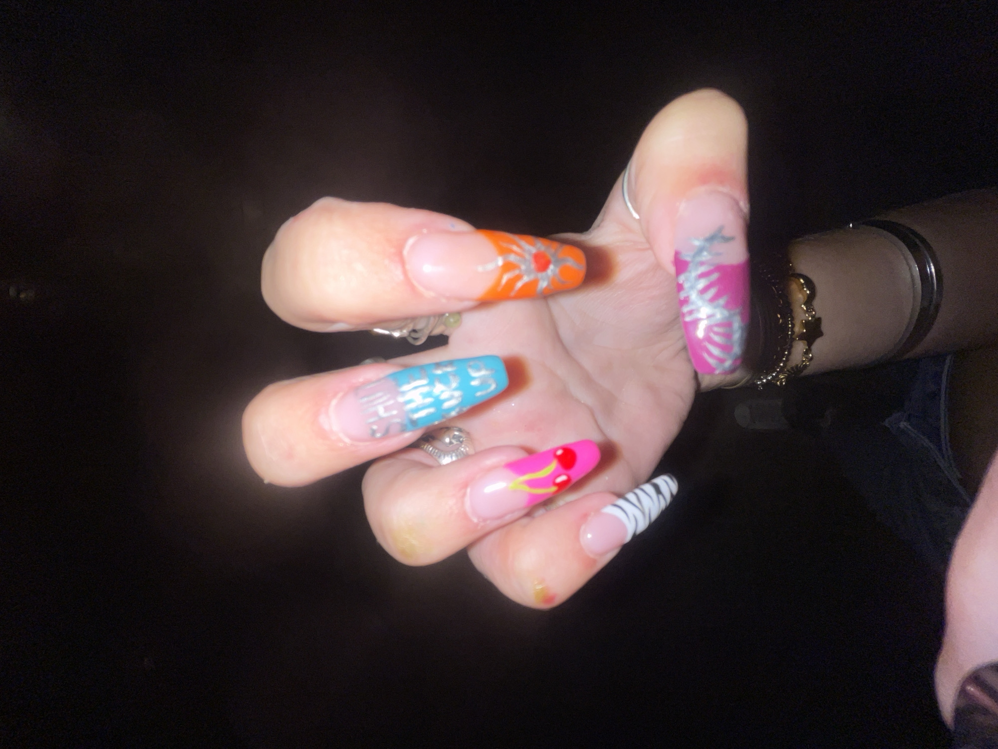 A person's hand with long, artificial nails is shown against a dark background. The nails are decorated with various designs. The index finger has a pink base with a yellow stem and two red cherries. The middle finger is a gradient of pink to blue with silver text that appears to read "SHUT THE FUCK UP." The ring finger has an orange base with a silver sun design. The little finger has a pink base with a silver design of what appears to be barbed wire. The thumb has a pink and white zebra stripe pattern. Several rings are visible on the fingers, and bracelets are worn on the wrist. The background is predominantly black, suggesting an indoor or nighttime setting. There is no discernible indication of the location being Raanana, Israel.