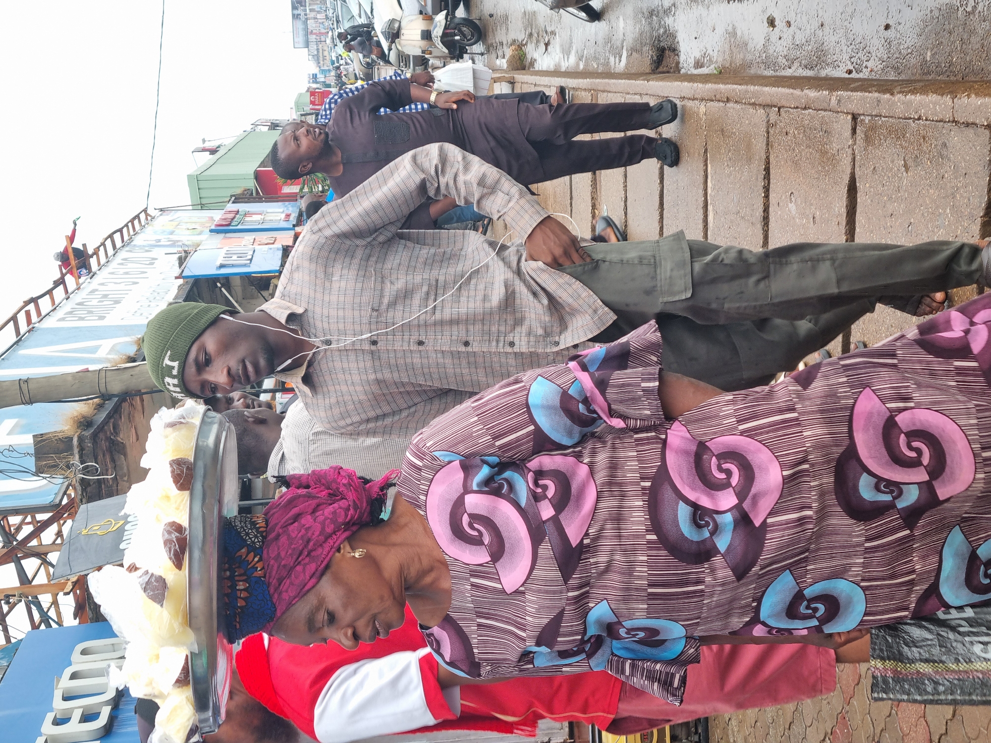 In Jos, Nigeria, the scene captures a busy street with several individuals present. An elderly woman is in the foreground, wearing a red top and a patterned wrapper with pink and blue spiral designs. She's carrying a large tray on her head, filled with what appears to be food items. A younger man with a green beanie and wearing a plaid shirt stands next to her. He has earphones in. Behind them, another man is seen walking with his hand in his pocket. He is wearing a brown outfit. The background includes buildings with signs, scooters, and other elements typical of a commercial street. The street appears wet, suggesting recent rain.