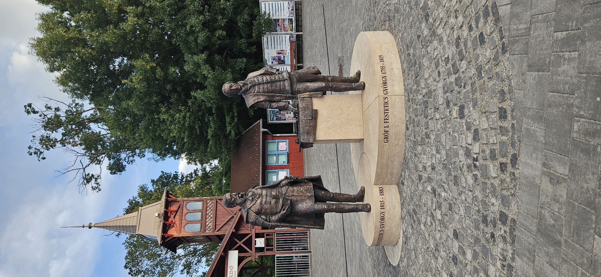 The image displays a bronze statue of two men in historical attire standing on a circular stone pedestal in Hévíz, Hungary. The statue is situated in an outdoor public space with cobblestone paving in the foreground. Behind the statue, there is a red wooden building with a distinctive tower and a tiled roof, suggesting it might be part of a historical or tourist-oriented site. Large green trees form a natural backdrop, and the sky is partly cloudy. The inscription on the pedestal identifies the figures as "GRÓF I. FESTETICS GYÖRGY 1755 - 1819" and "GRÓF GYÖRGY FESTETICS 1815 - 1883". This memorial honors members of the Festetics family, who were influential landowners in the region. The overall scene conveys a sense of historical significance and public commemoration.