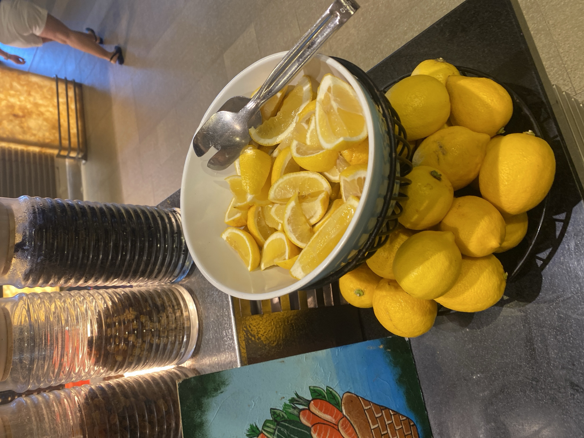 The image shows a close-up view of a buffet selection featuring lemons. In the foreground, a black basket overflows with whole, bright yellow lemons. Above this, a white bowl contains a generous portion of sliced lemons, with a silver serving utensil resting on top. To the left of the lemons is a colorful, painted board depicting a basket of carrots. In the background, three tall glass jars filled with what appear to be dried goods are visible. The overall setting suggests a food service area, possibly in a hotel or restaurant. There are no people visible in the immediate foreground. The location is not specified in the image.