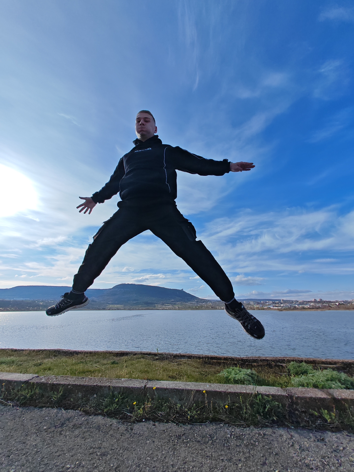 A full-length shot captures a person mid-air, seemingly jumping, with a backdrop of a landscape featuring water, mountains, and a city skyline. The person, dressed in a black tracksuit, appears to be in motion. Their arms are extended out to their sides, and legs are spread apart in a jumping pose.
The background consists of a body of water, likely a lake, with mountains in the distance. On the far right, a city skyline is visible. The sky is bright blue with wispy clouds, indicating a clear day. The location is near a body of water in the city of Shumen, Bulgaria.