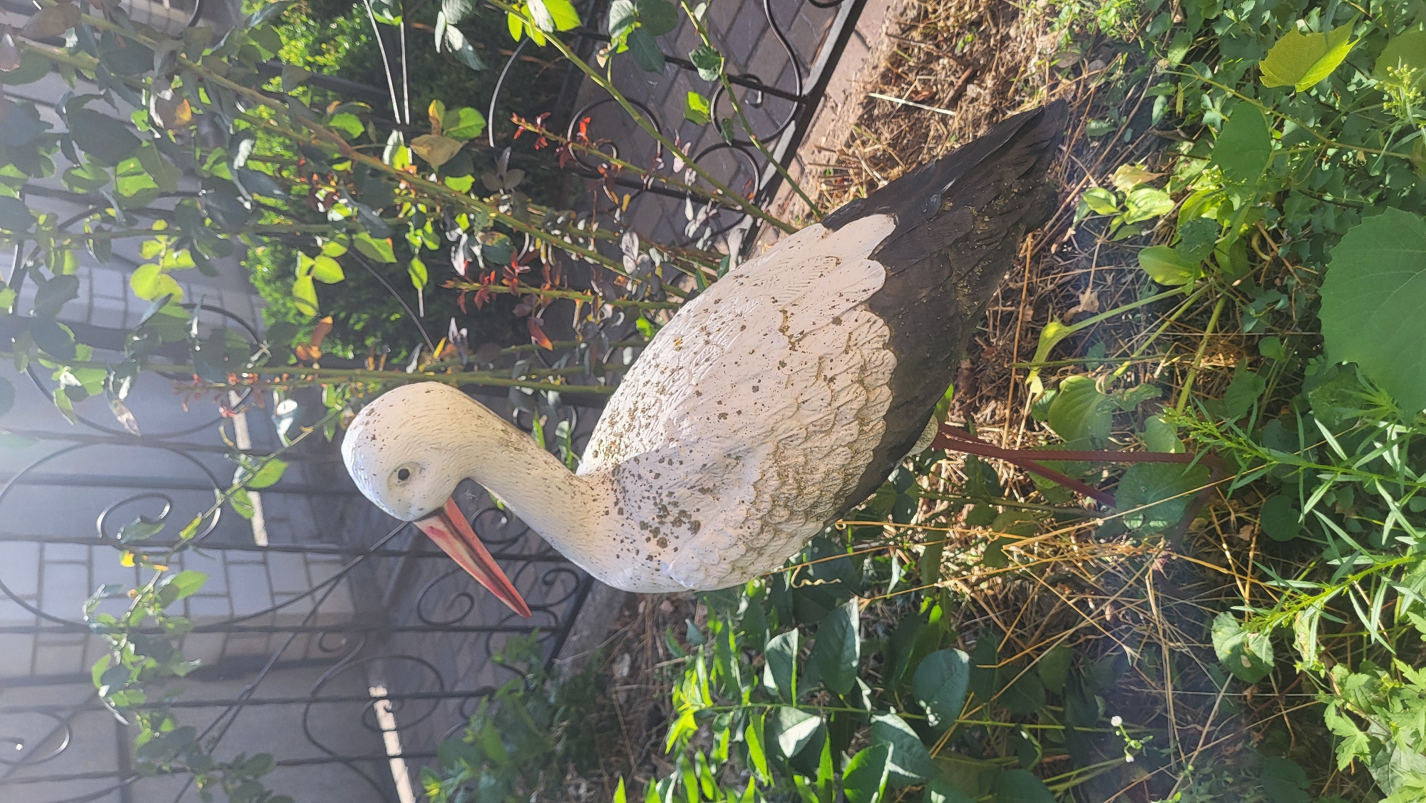A decorative garden statue of a stork stands in a garden. The stork is white with black wings and legs, and a red beak. It is standing on one leg, with its other leg tucked underneath its body. The statue is in focus and takes up most of the frame. The background is blurred and shows green foliage and a black metal fence. The lighting suggests it is daytime, with sunlight casting shadows. The scene appears to be outdoors in a residential or park setting.
