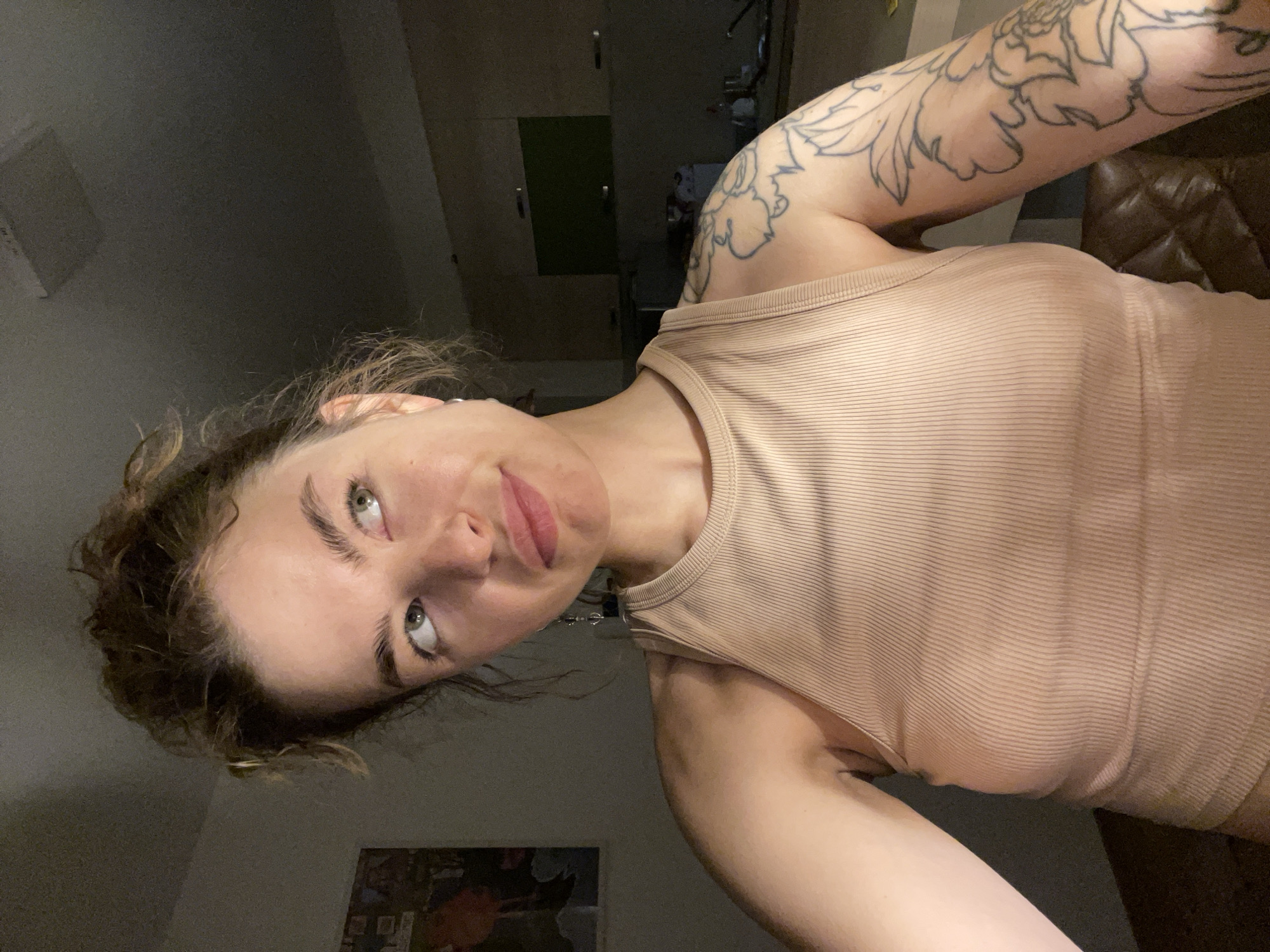 A young woman with fair skin and curly hair poses indoors, wearing a beige ribbed tank top. Her left arm, prominently displayed, features a large floral tattoo. She is tilting her head and looking up with a slight smirk. The background includes a kitchen area with cabinets, a leather chair, and a picture on the wall. The scene appears to be dimly lit. There is no explicit indication the photo was taken in Boreti, Montenegro.