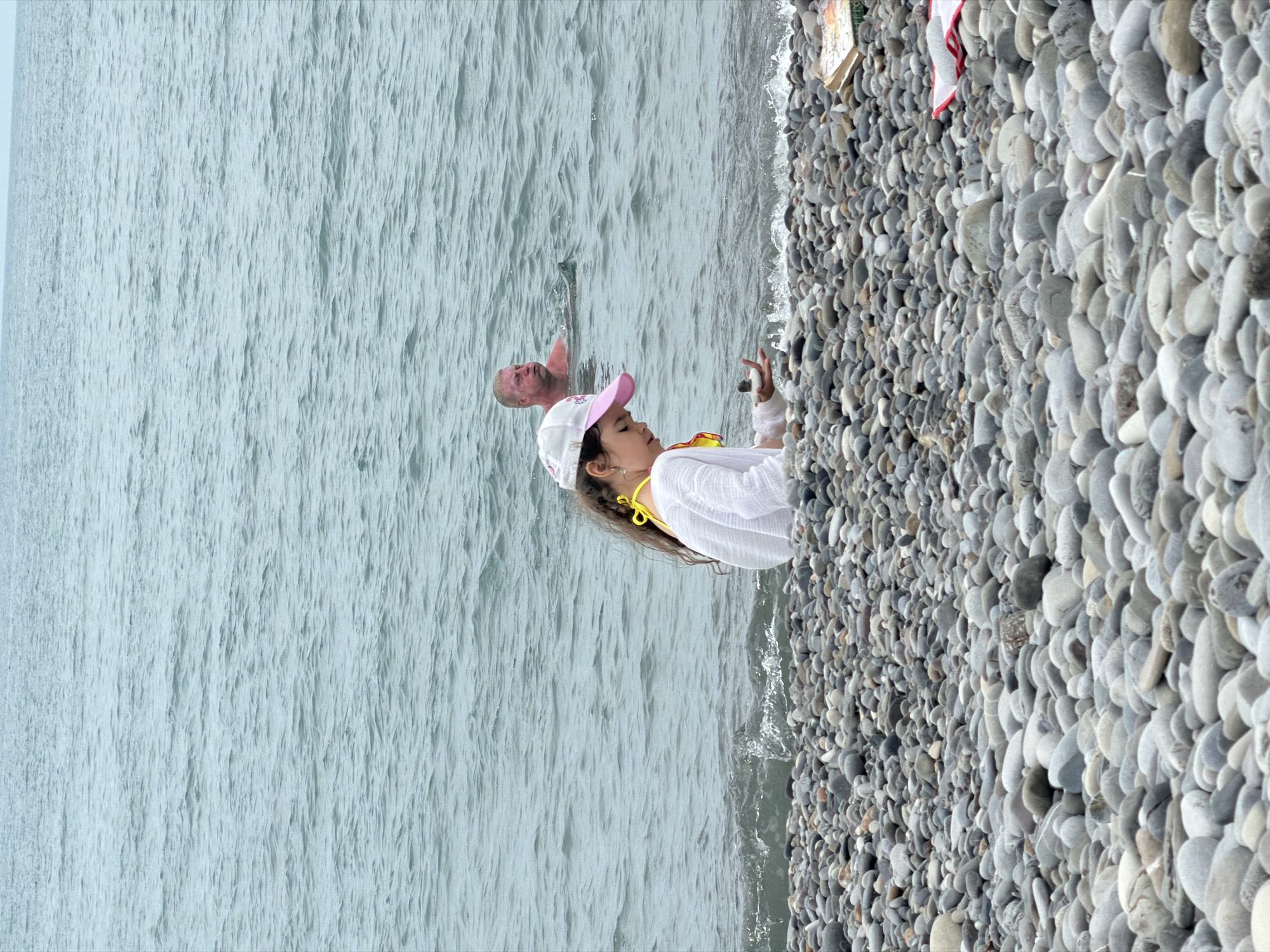 A medium shot shows a pebble beach and the sea. A young girl wearing a white shirt, yellow bathing suit, and a pink and white baseball cap is sitting on the beach, holding a small stone in her hand. In the background, a man is swimming in the sea. The water is choppy and grey. The beach is covered in smooth, grey pebbles of various sizes. A book and a red and white striped object are visible on the beach to the right. The scene appears to be a public beach in Sochi, Russia.