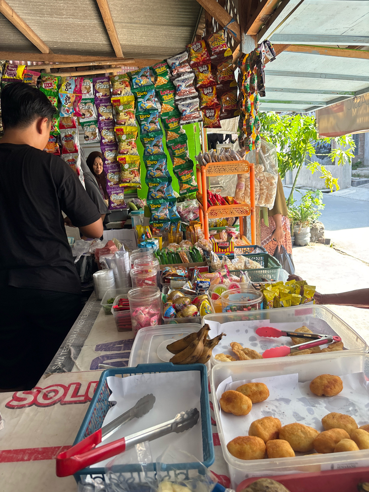 This image depicts a street-side food stall in Tegal, Indonesia. The stall is laden with a variety of packaged snacks and some freshly prepared food items. In the foreground, two clear plastic containers hold fried snacks. The container on the right contains numerous small, golden-brown fried dough balls, while the container to its left holds what appear to be fried plantain slices and other assorted fried items. A bunch of bananas is also visible in the upper left corner of this container. Two pairs of tongs, one with red handles and the other silver, are placed on top of the snacks, suggesting they are for serving.
Behind the food display, a person wearing a black t-shirt and black pants stands facing away from the camera, appearing to be a customer. To their left, a woman wearing a hijab and a patterned garment is partially visible, looking towards the snacks. A rack filled with various brands of Indonesian snack bags, such as "Garuda Pilus" and "Taro Net," is prominently displayed behind them. Additional items like bottled drinks, plastic cups, and small packets of food are also on display on shelves and counters. The stall is covered by a corrugated metal roof supported by wooden beams. The background shows a slightly blurred outdoor environment with some greenery and a street. The overall scene captures a typical small business operation in a local Indonesian setting.