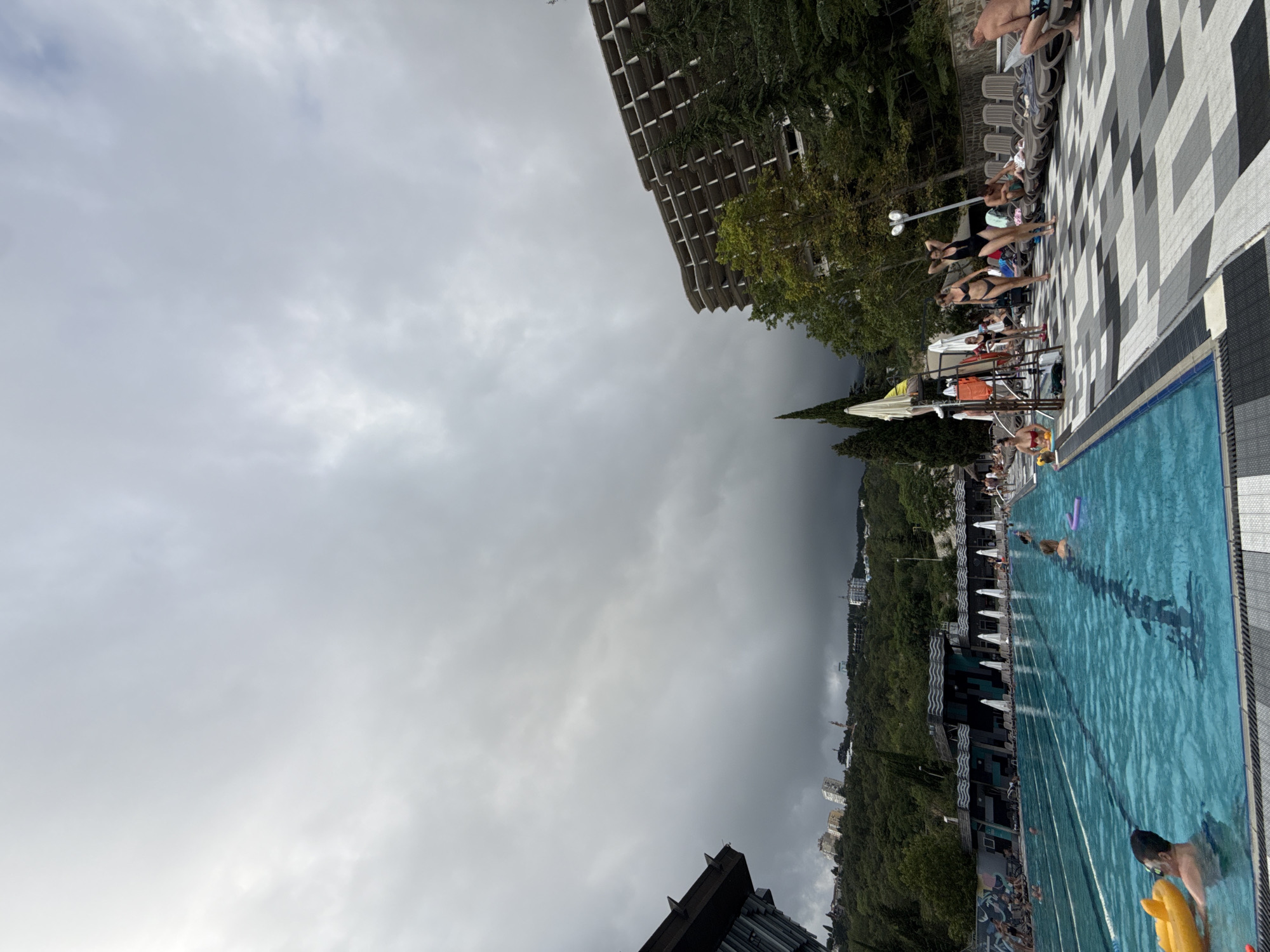 The image depicts a large outdoor swimming pool complex with a group of people enjoying the water and the surrounding amenities. The pool itself is a vibrant blue, with lane markers visible, indicating it is designed for lap swimming as well as recreational use. Several individuals are in the water, some swimming, others floating on inflatable toys. Along the edge of the pool, lounge chairs are set up, with some people sunbathing or relaxing. A building with a modern architectural design, featuring numerous balconies and concrete structures, dominates the background on the right side of the image. This building appears to be either under construction or in a state of disrepair. To the left, another building with a darker, more angular facade is visible. The entire scene is set against a backdrop of lush green hills covered with trees, with several buildings of varying sizes scattered across the slopes, suggesting an urban environment nestled within a natural landscape. The sky above is overcast with heavy, dark clouds, hinting at potentially stormy weather. The location is identified as a swimming pool in the city of Yalta, Ukraine.