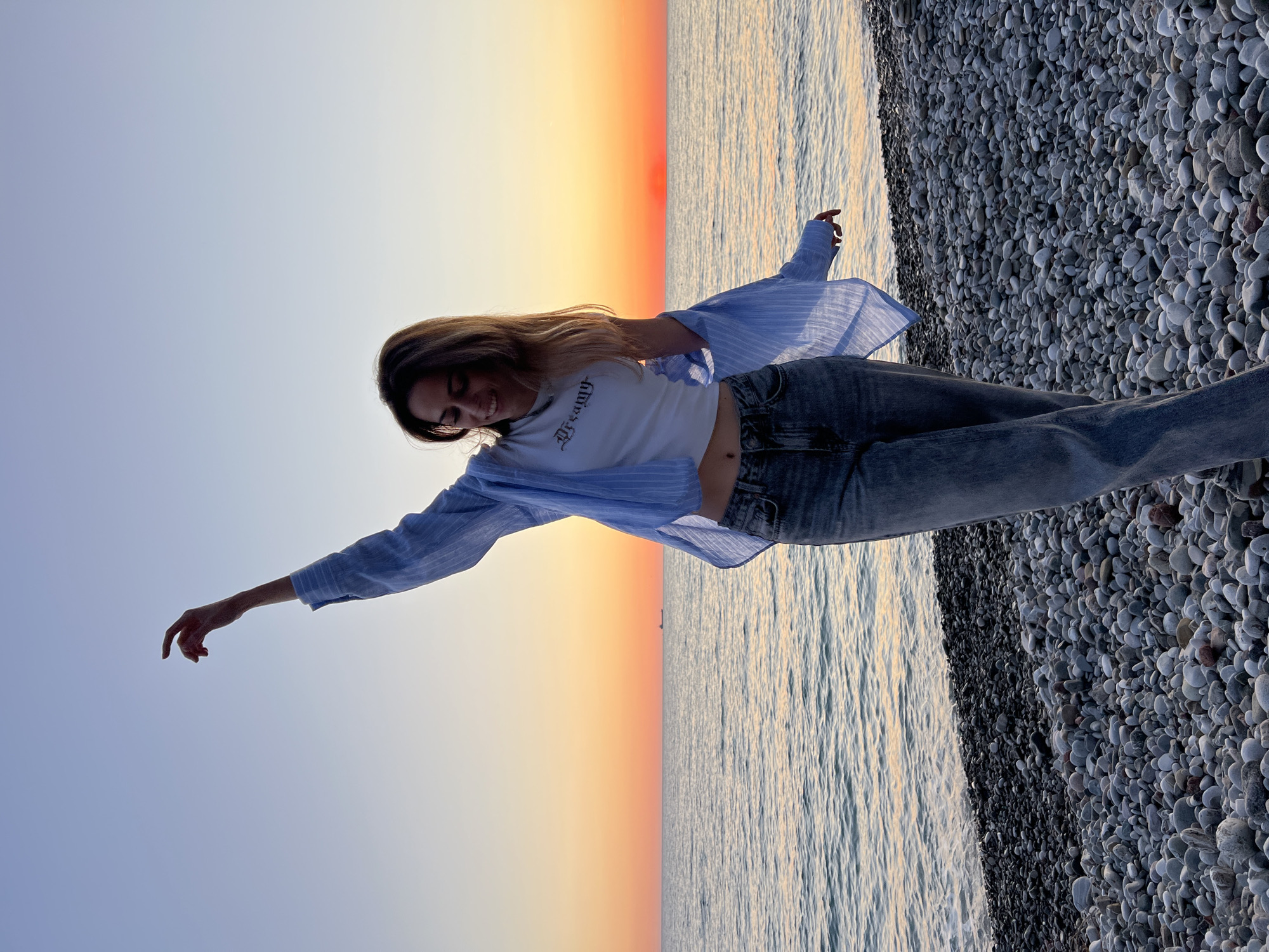 A young woman stands on a rocky beach at the edge of the sea, likely in or near Bar, Montenegro, given the pebble beaches characteristic of the region. She is wearing a white t-shirt with the word "dreamy" printed on it, a light blue striped shirt draped open, and blue jeans. She is smiling and extending her arms outward. The sky is a gradient of colors, from a soft blue to shades of orange and red as the sun sets or rises. The water reflects the light. The beach is covered in smooth, gray pebbles.