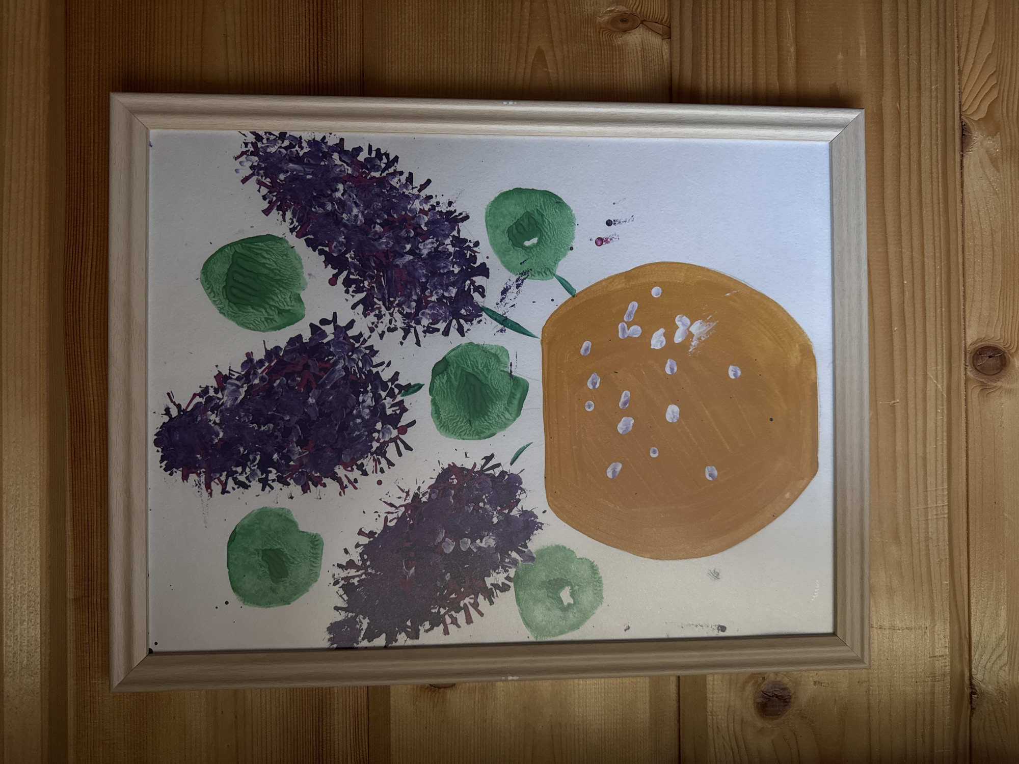 A framed child's artwork hangs on a wooden wall. The artwork depicts purple flower clusters, green circular shapes, and a tan, round form with white dots. The wooden wall has vertical planks. The location is unspecified.