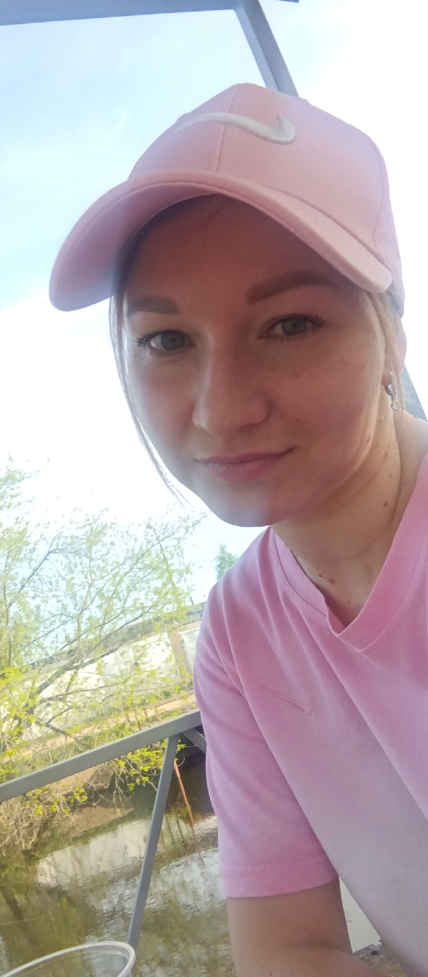 A woman with light skin and blond hair is wearing a pink Nike baseball cap and a pink shirt. She is standing outside on what appears to be a balcony or bridge, with a body of water visible below. A metal railing is present, and trees with budding green leaves are visible in the background. The sky is partly cloudy. The location is not identifiable from the image.