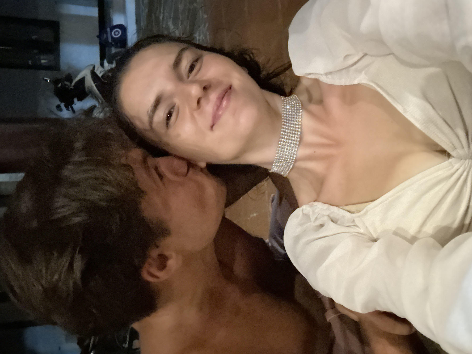A fair-skinned woman with dark hair, wearing a white top with a diamond choker necklace, is being kissed on the cheek by a light-skinned male with short, dark brown hair. They are indoors, and the background is blurry. There is a partially visible black object in the background. The exact location within Mũi Né, Vietnam cannot be determined from the image.