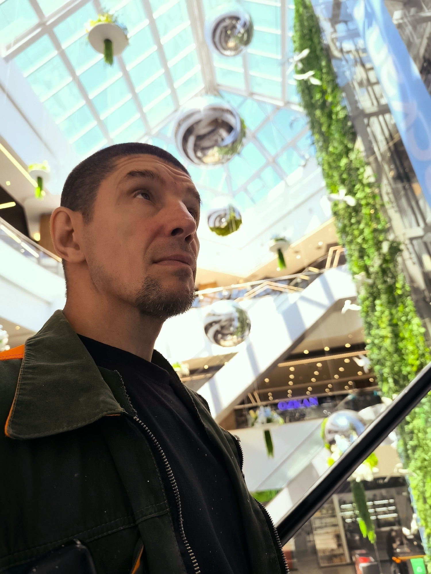 This media file is a close-up, low-angle shot of a man looking upwards in what appears to be a modern, multi-story indoor space, likely a shopping mall or atrium. The man is positioned in the foreground, on the left side of the frame, wearing a dark jacket with an orange trim and a black t-shirt underneath. He has short, dark hair and a goatee. His gaze is directed upwards and slightly to the right, with a thoughtful or contemplative expression.
The background is blurred but reveals several notable features of the environment. The ceiling is a large, transparent or translucent structure, allowing natural light to filter through, suggesting it is daytime. Suspended from the ceiling are several large, spherical, reflective decorations, reminiscent of disco balls, and some hanging green floral or plant arrangements. Multiple levels of balconies or walkways are visible, with railings and escalators. A prominent feature on the right side of the frame is a tall, vertical installation covered in lush green plants, creating an indoor garden effect. There are also decorative white bird-like figures hanging on the greenery.
The overall impression is one of a bright, modern, and well-decorated interior. The activity taking place is not explicit, but the man's upward gaze and the environment suggest he might be observing the architecture, the decorations, or simply enjoying the atmosphere. The lighting suggests it is daytime. There is no discernible weather information.
A small piece of text is visible in the background, blurred but appears to say "OQAN" in blue letters on a white banner or sign.
The location context provided is Qarağandı city administration, Kazakhstan, which might suggest this is a public building or a place of commercial activity within that city.