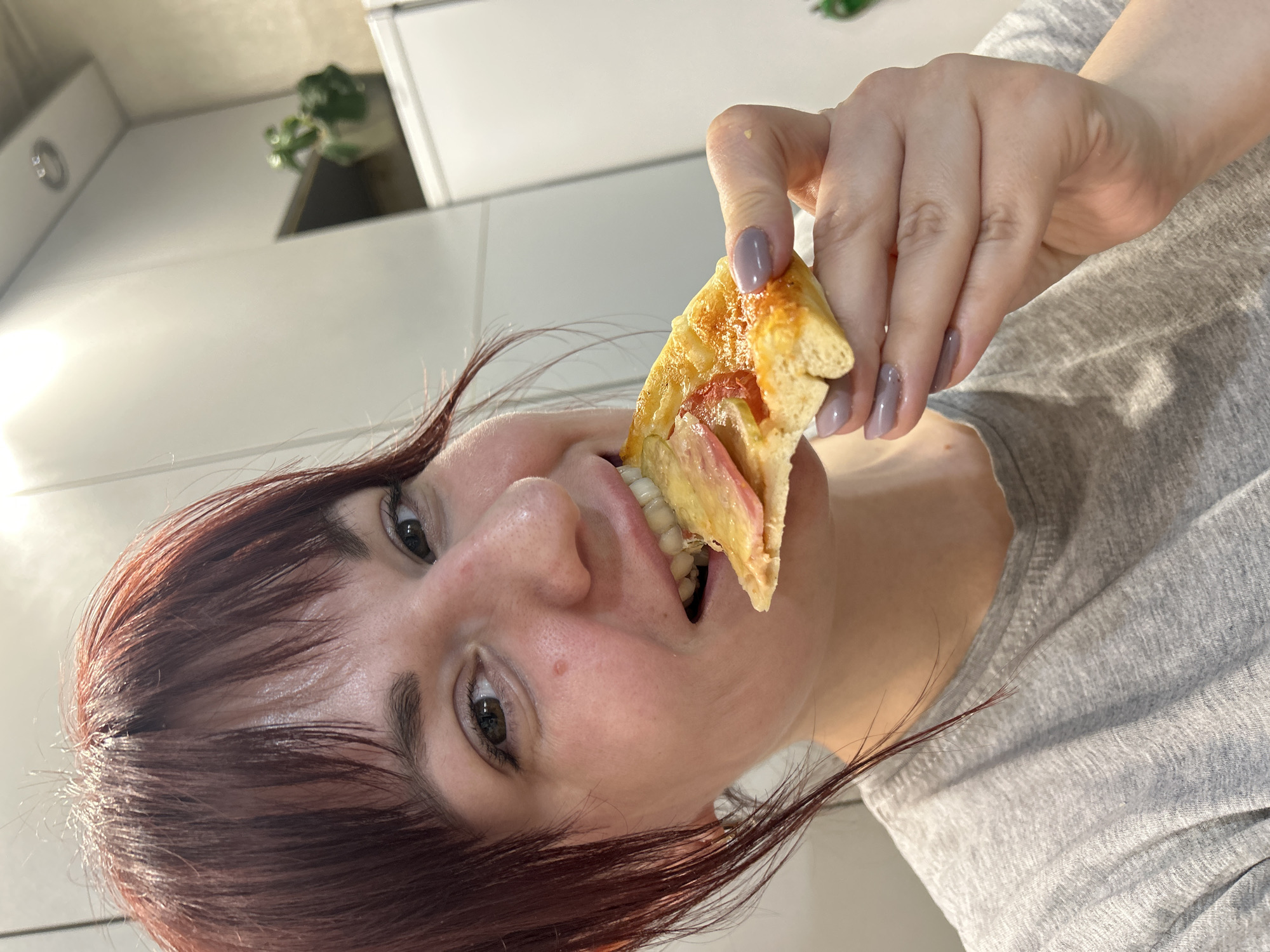 A woman with auburn-colored hair is eating a piece of pizza indoors. The woman is wearing a gray t-shirt and has light skin. Her fingernails are painted light purple. The pizza appears to have cheese, tomatoes, and ham. The background includes white cabinets and a small potted plant. The specific location in Фоки, Russia is not evident from the image.