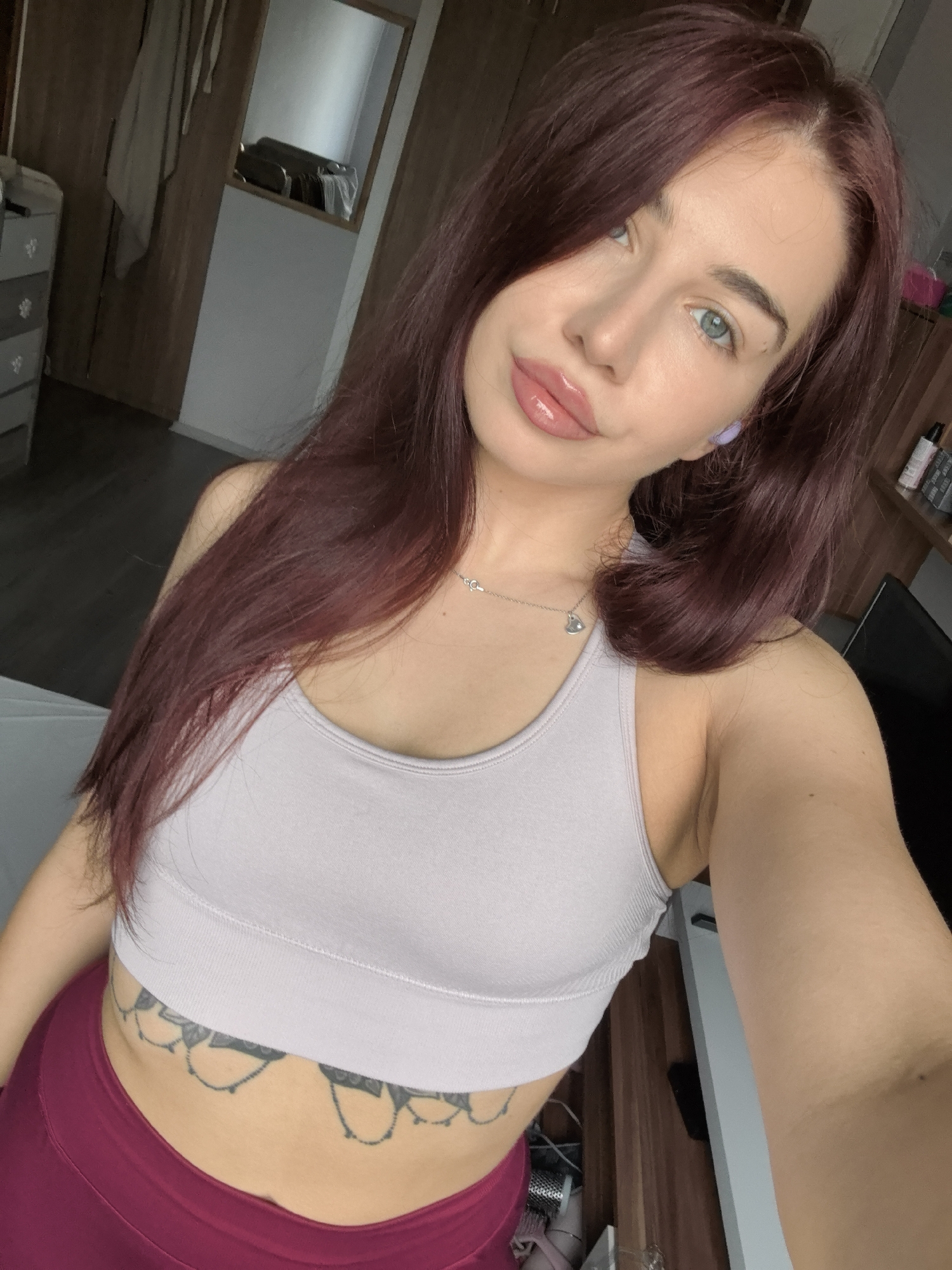 A young woman with long, dark reddish-brown hair is captured in a selfie-style shot, looking directly at the camera with a slight smile. Her hair is parted and falls over her left shoulder and down her back. She has light skin, blue-green eyes, defined eyebrows, and full lips accented with gloss. She wears a light grey ribbed sports bra and high-waisted dark pink or magenta leggings. A silver necklace with a heart-shaped pendant hangs around her neck, and a small purple earbud is visible in her right ear. A decorative, mandala-like tattoo design is visible across her lower abdomen, just below the sports bra.
The setting appears to be an indoor room, likely a bedroom or personal space, with natural light suggesting daytime. In the background to her left, a wooden wardrobe or cabinet and a grey dresser with decorative handles are visible. A mirror above the dresser reflects part of a window or light source. To her right, shelves hold various bottles and containers, and the edge of a dark chair or piece of furniture is present. No text is visible within the image. The location context provided is Stara Zagora, Bulgaria.