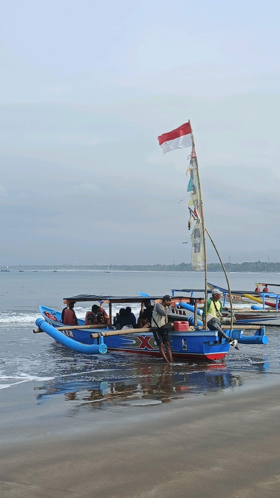 A boat with the Indonesian flag is partially submerged in shallow water at the beach in Desa Pagandaran, Indonesia. Several people are visible on the boat, some standing and some sitting. One person is standing near the rear of the boat and another is sitting near the front. The boat is blue with red and white accents and appears to be a fishing boat. The water is calm and the sky is overcast. In the background, there are other boats and a distant shoreline with trees. The sand of the beach is visible in the foreground.